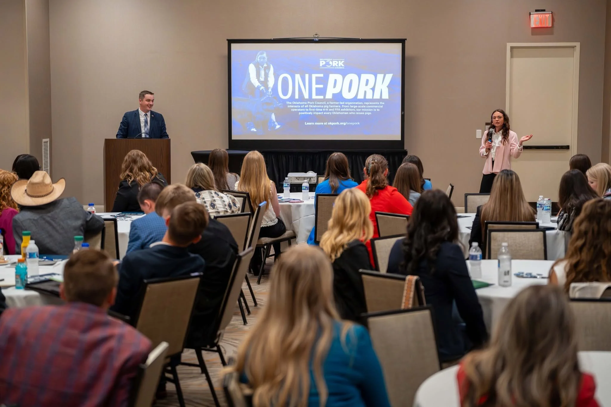 A conference room with people sitting at round tables, listening to a presentation. Two speakers, a man at a podium and a woman standing with a microphone, present in front of a large screen displaying 'ONE PORK' and informational text about the Oklahoma Pork Council. The audience includes men and women of various ages, some with water bottles and notebooks on the tables.