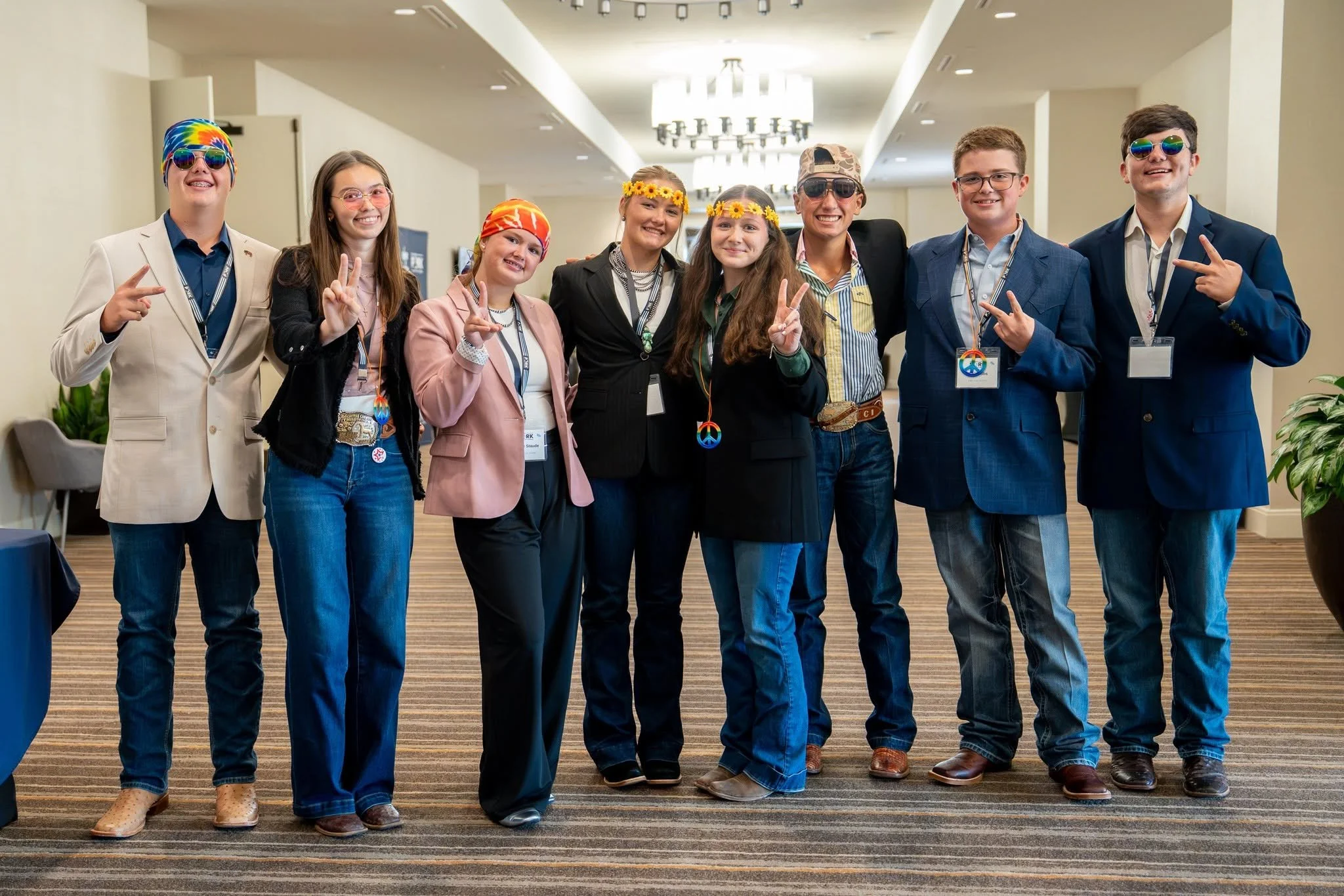 Nine young adults standing together in a brightly lit hotel lobby, smiling, wearing colorful accessories, some with peace sign hand gestures, dressed in casual and semi-formal attire, posing for a group photo.