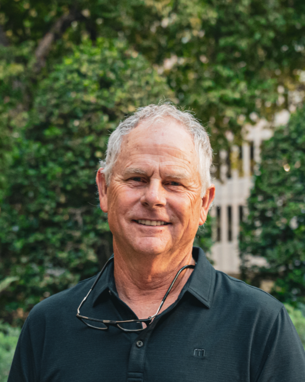 A smiling older man wearing a light gray baseball cap with "DAD" embroidered on it and a white T-shirt with a small bunny logo, standing outdoors with a blurred background.