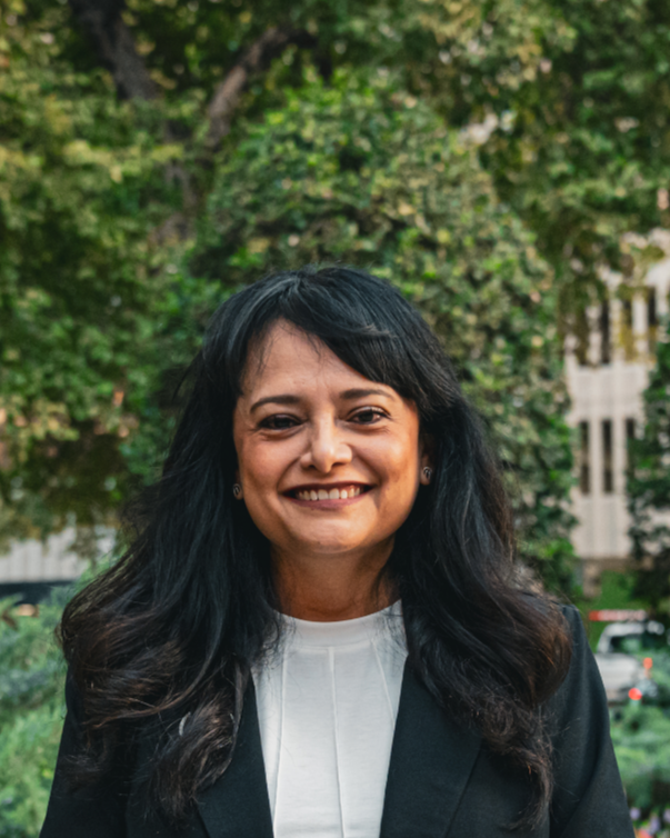 Portrait of a woman with shoulder-length dark hair, wearing a dark blazer and light blue shirt, smiling against a plain white background.