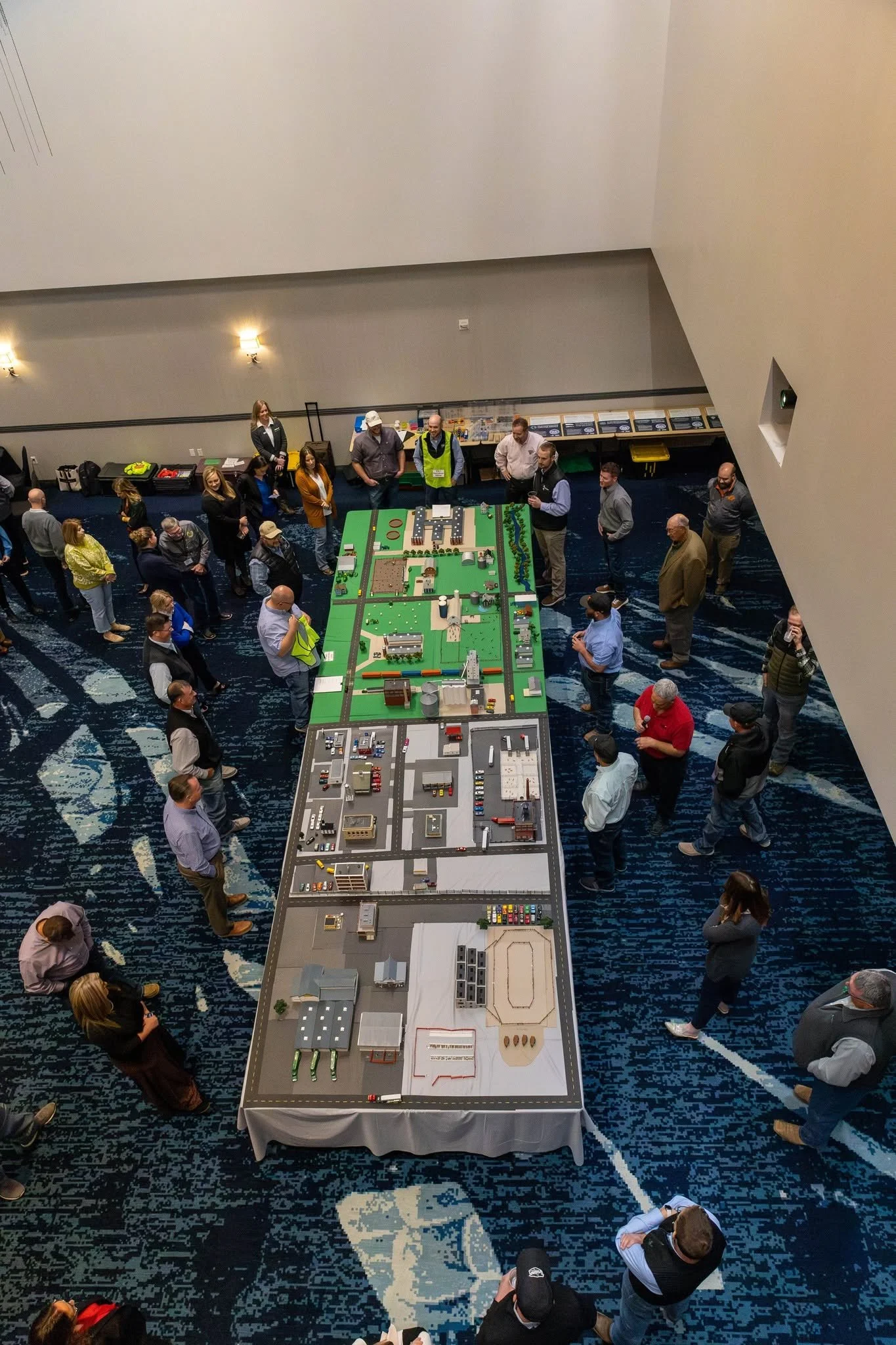 A large model city layout on a long table with miniature buildings, roads, and infrastructure. People are gathered around observing and discussing the display in a conference or exhibit setting.