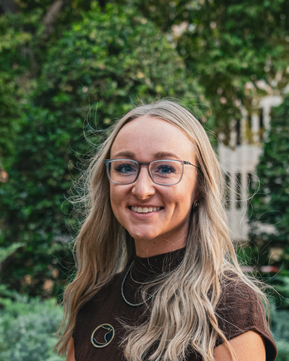 A woman with long blonde hair, glasses, and a white striped shirt smiling outdoors with greenery in the background.