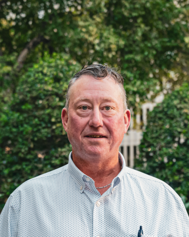 A man with short gray hair wearing a black Oklahoma Pork Council vest, white shirt, and tie, standing outdoors in front of a blurred background of trees and modern buildings.