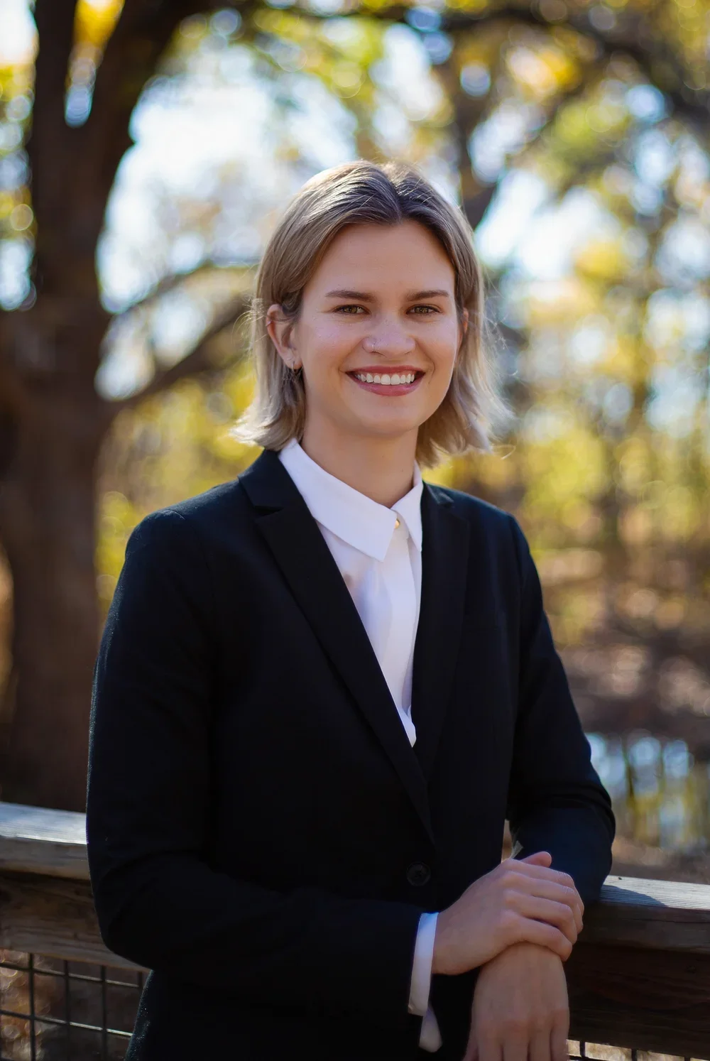 A young woman with shoulder-length blonde hair, smiling, in a black blazer and white shirt, outdoors during daytime with trees and blurred autumn leaves in the background.