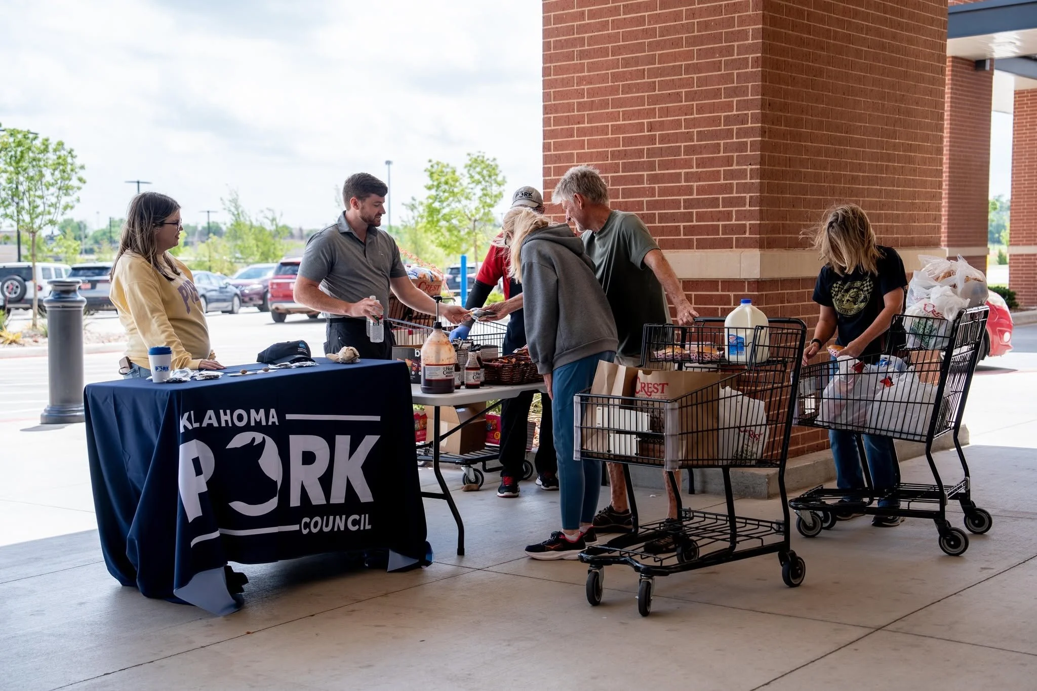 People shopping at a grocery donation event outside a building with a brick wall. A table displays flyers and donations, with a sign reading 'OKLAHOMA PARK COUNCIL.' Customers are picking up groceries from a table, with shopping carts nearby.