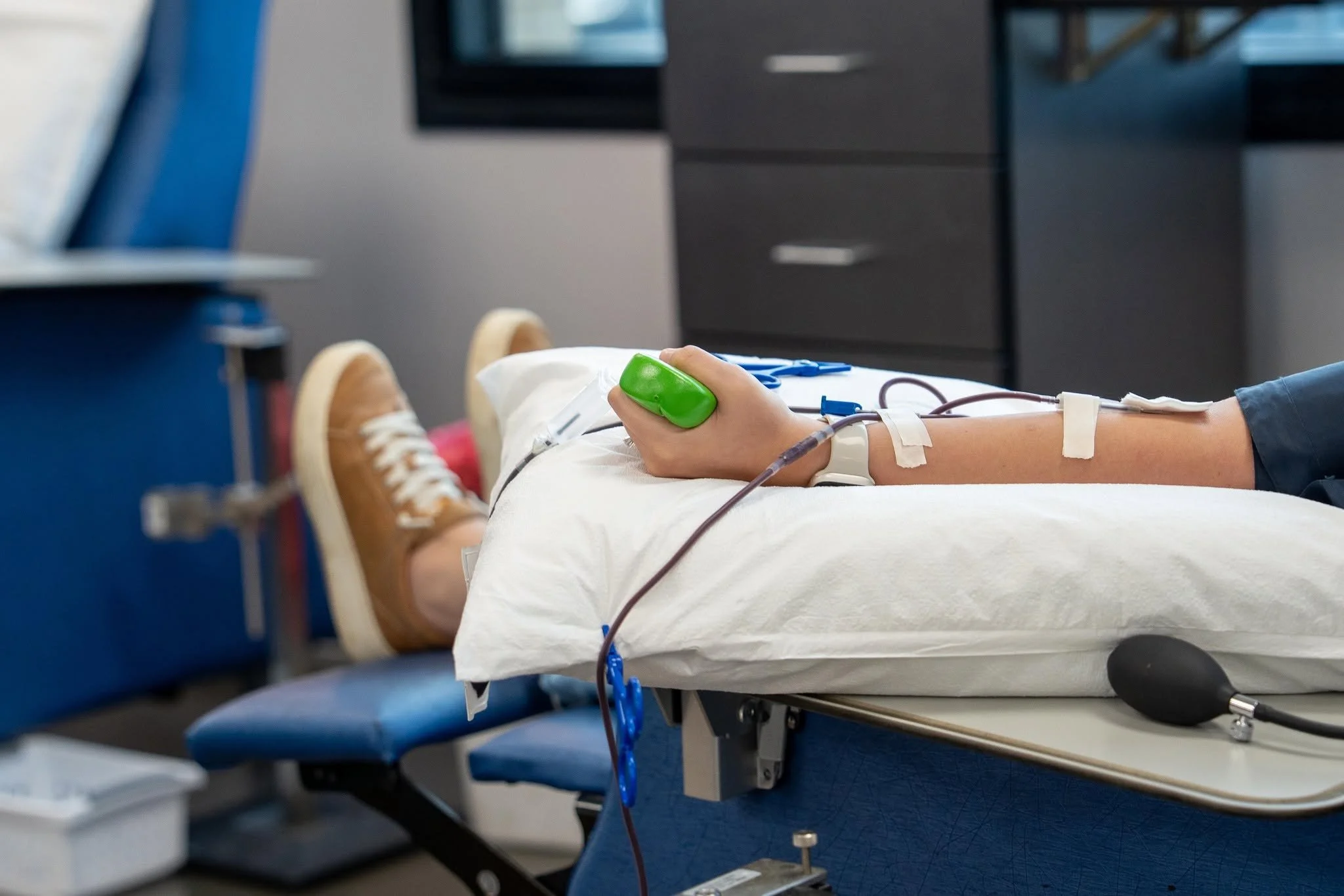 A person lying on a hospital bed with a blood pressure cuff on their arm, connected to medical tubing and equipment, in a healthcare setting.