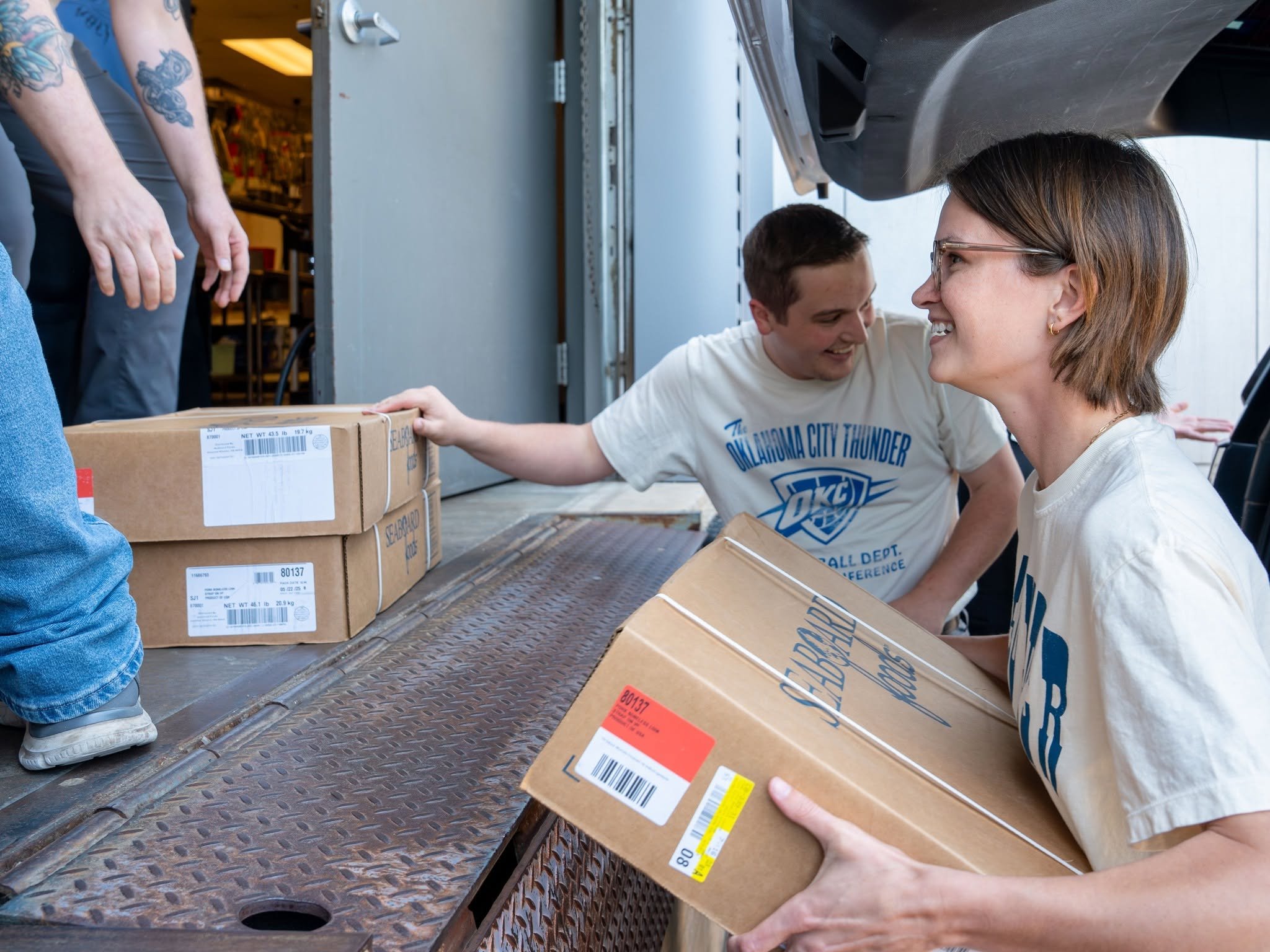 People exchanging boxes at a loading dock. One person is handing a cardboard box to another person who is smiling. The boxes have labels and barcodes on them. The scene appears to be in a warehouse or delivery area.