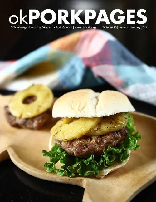 Cover of Oklahoma Pork Pages featuring a close-up of a cheeseburger with lettuce, cooked pineapple ring, and beef patty on a bun, served on a wooden tray, with a blurred background of a colorful cloth and a second burger in the background.