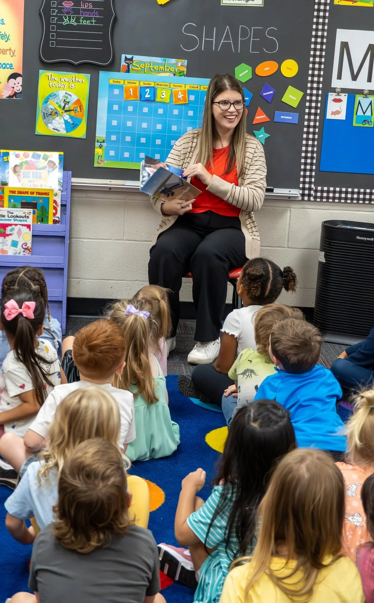 A woman with glasses, wearing a beige striped blazer and red shirt, sitting on a chair in front of a classroom filled with children gathering on a colorful rug. She is smiling and holding books. The blackboard behind her displays the word 'SHAPES' and has various educational posters and materials, including a weather chart and shape cutouts.