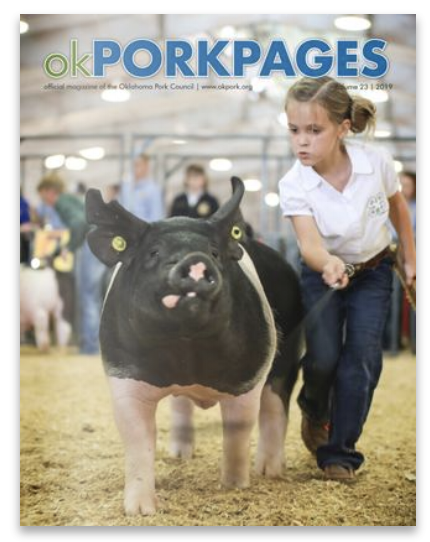 Cover of Oklahoma Pork Pages featuring a young girl guiding a black and white pig in a pig show at an indoor fair, with other participants and animals in the background.