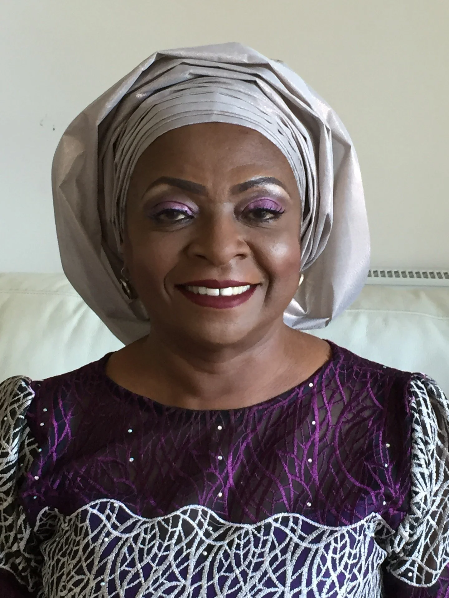 A smiling woman in a traditional African outfit with a large headwrap, purple and patterned dress, and makeup with pink eyeshadow and red lipstick, standing indoors against a light-colored wall.
