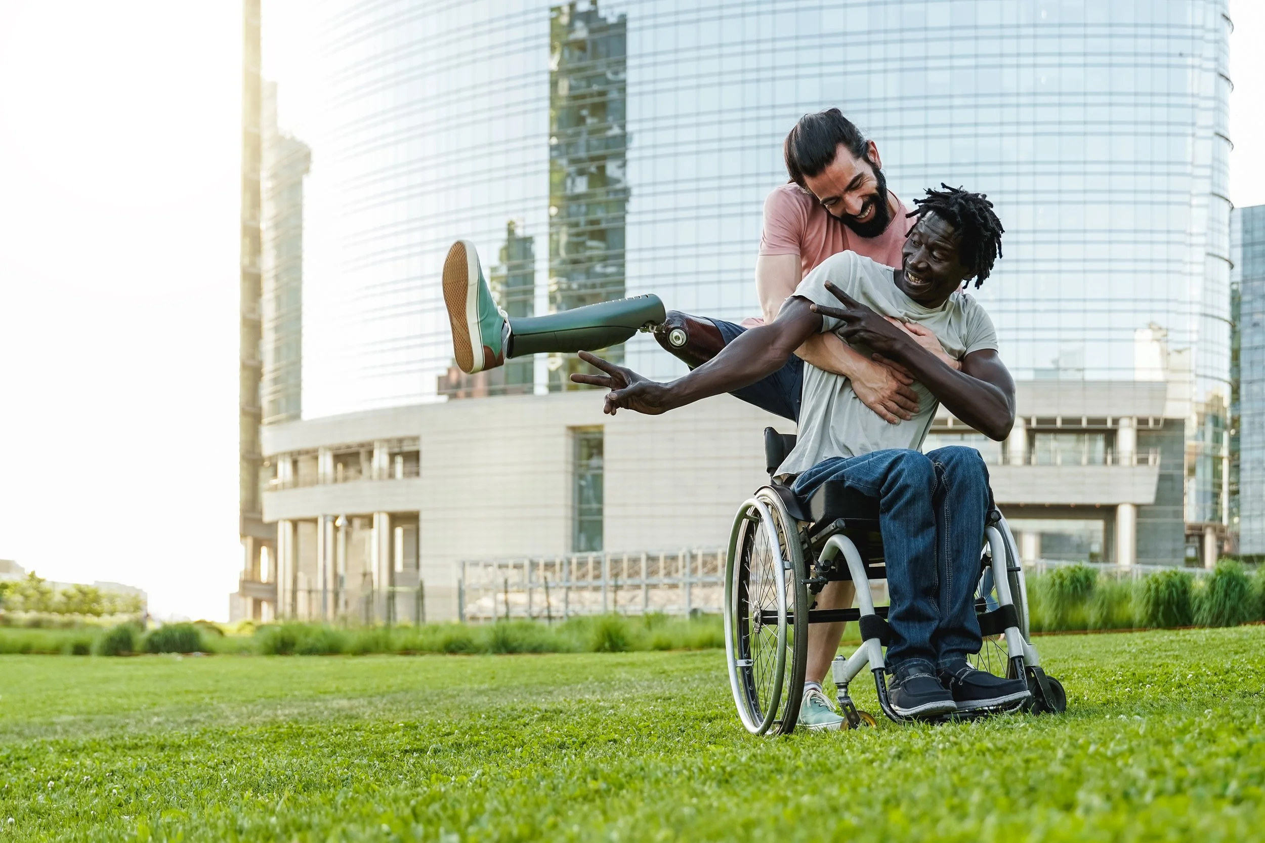 Two men in a park, one in a wheelchair, enjoying a playful moment near a modern building with reflective glass windows.