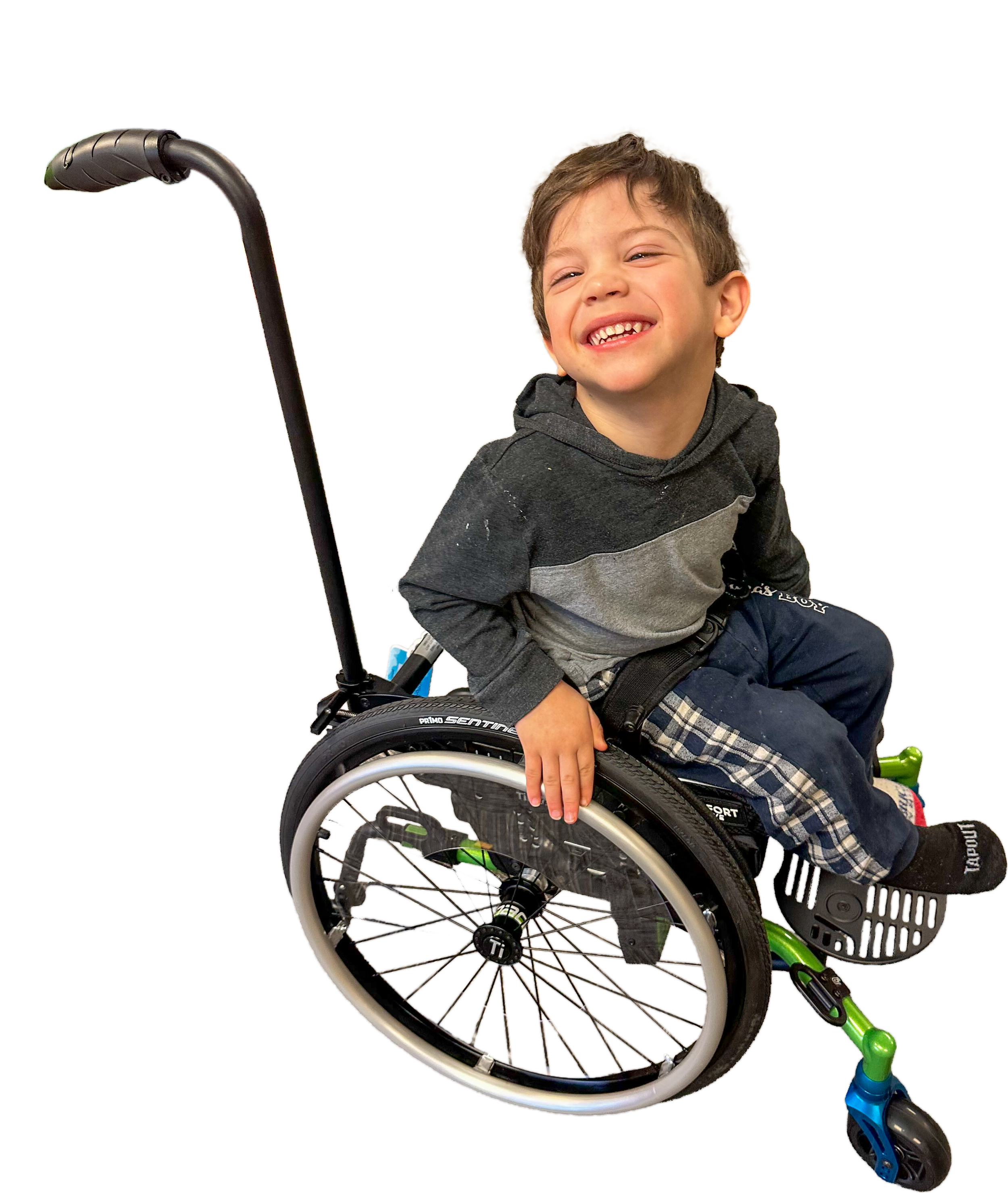 A young boy sitting in a wheelchair smiling with teeth showing, wearing a gray and black hoodie and plaid pants, with a scooter nearby.
