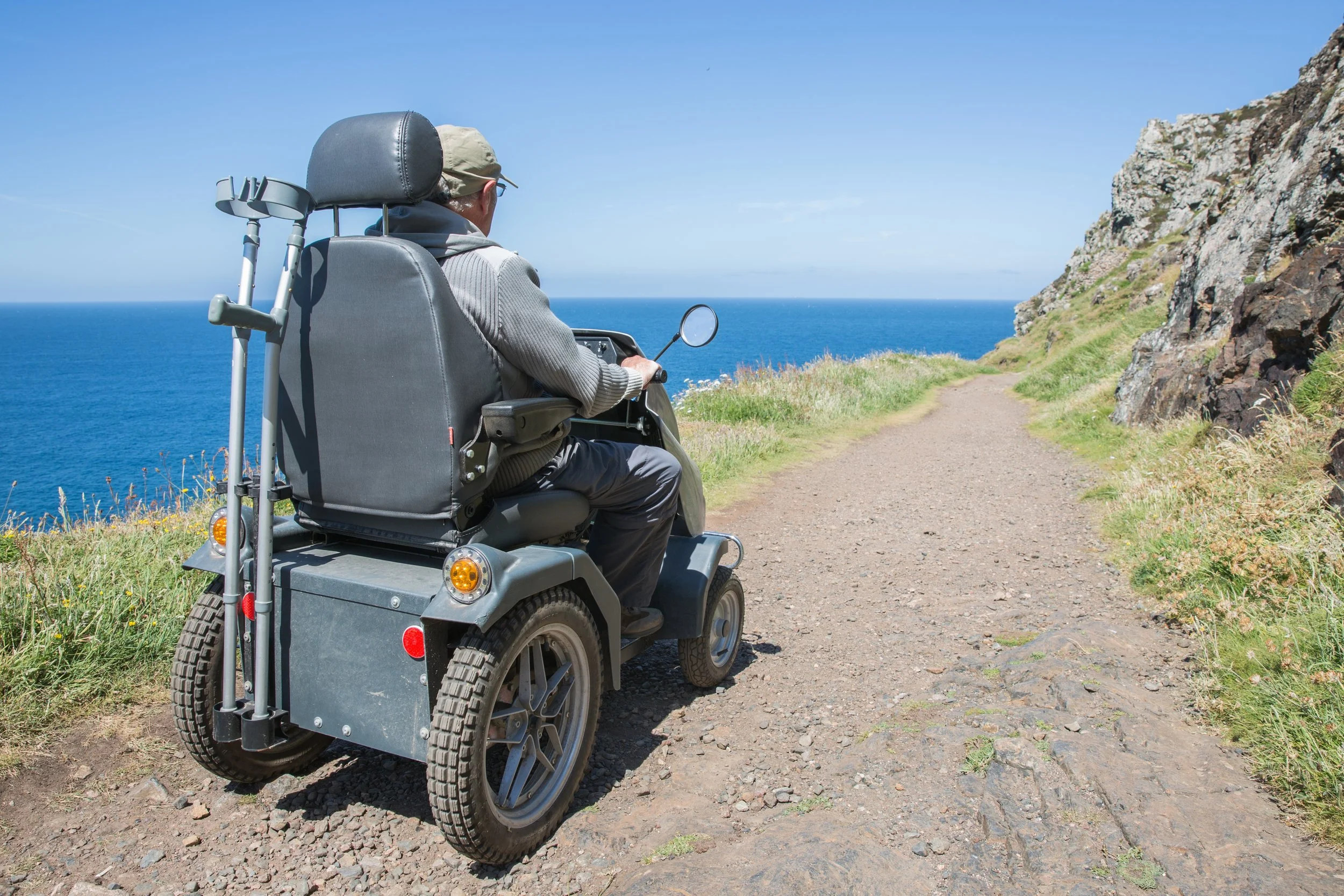 An elderly man in a mobility scooter driving along a dirt trail alongside a cliff, overlooking the ocean on a clear day.