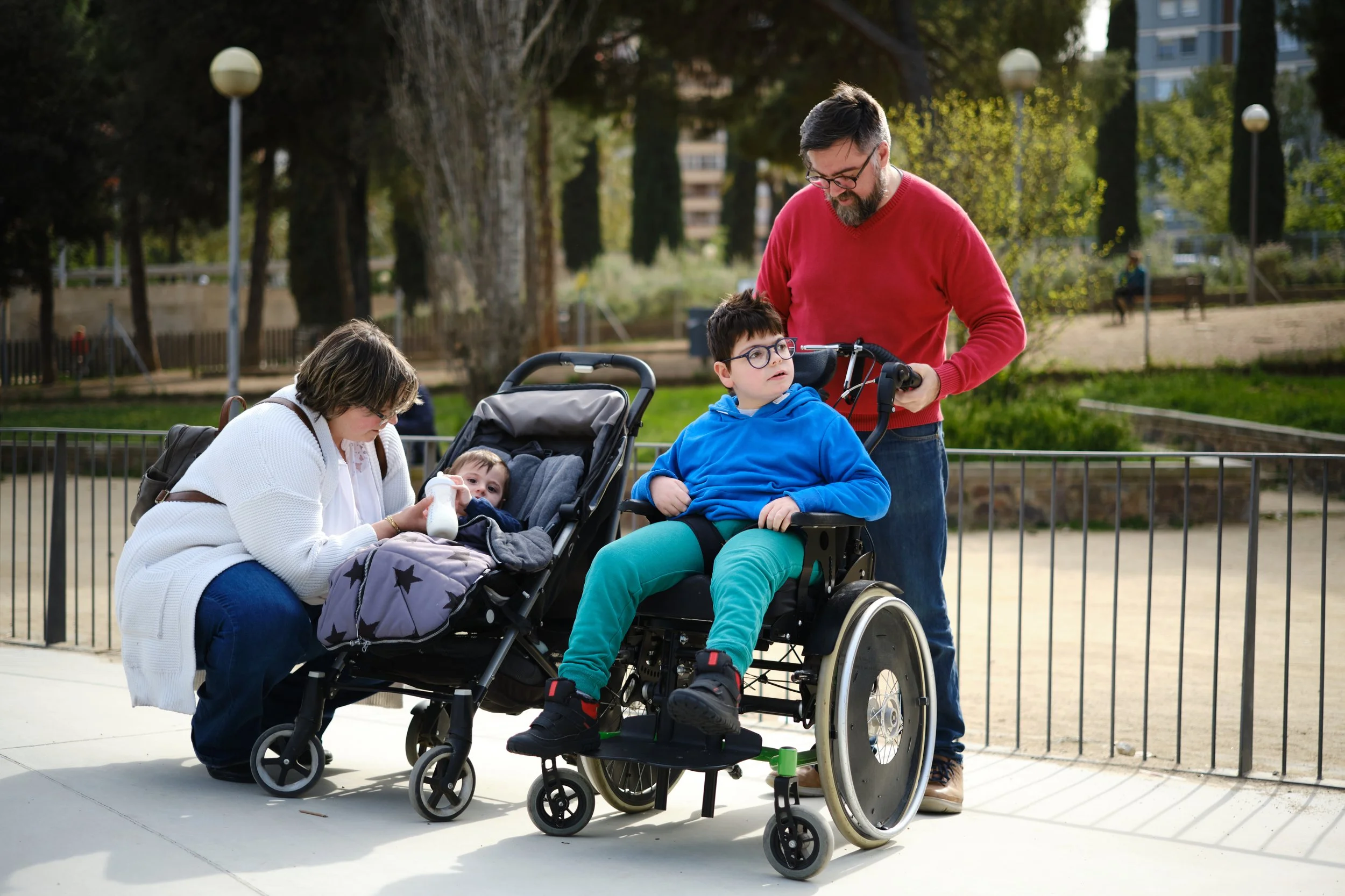 A man in a red sweater standing next to a little boy in a wheelchair, with a woman and a young child nearby in a park.