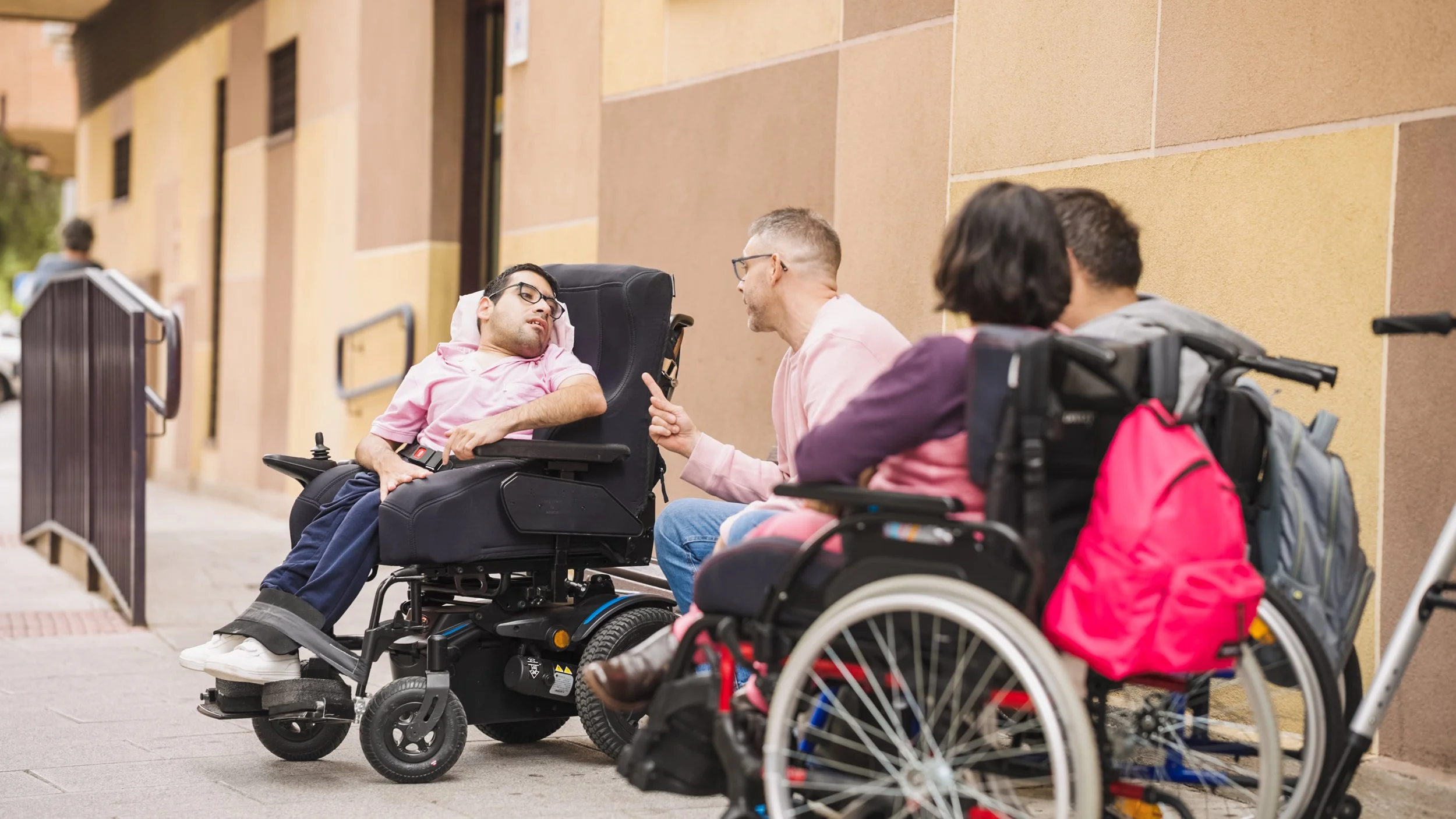 Four people sitting outside on a sidewalk, with two in wheelchairs and two on chairs. One person in a wheelchair is leaning back with eyes closed, while another person is talking to them, gesturing with a finger. The other two are sitting and facing away from the camera, with backpacks next to them. The scene is set against a yellow brick building.