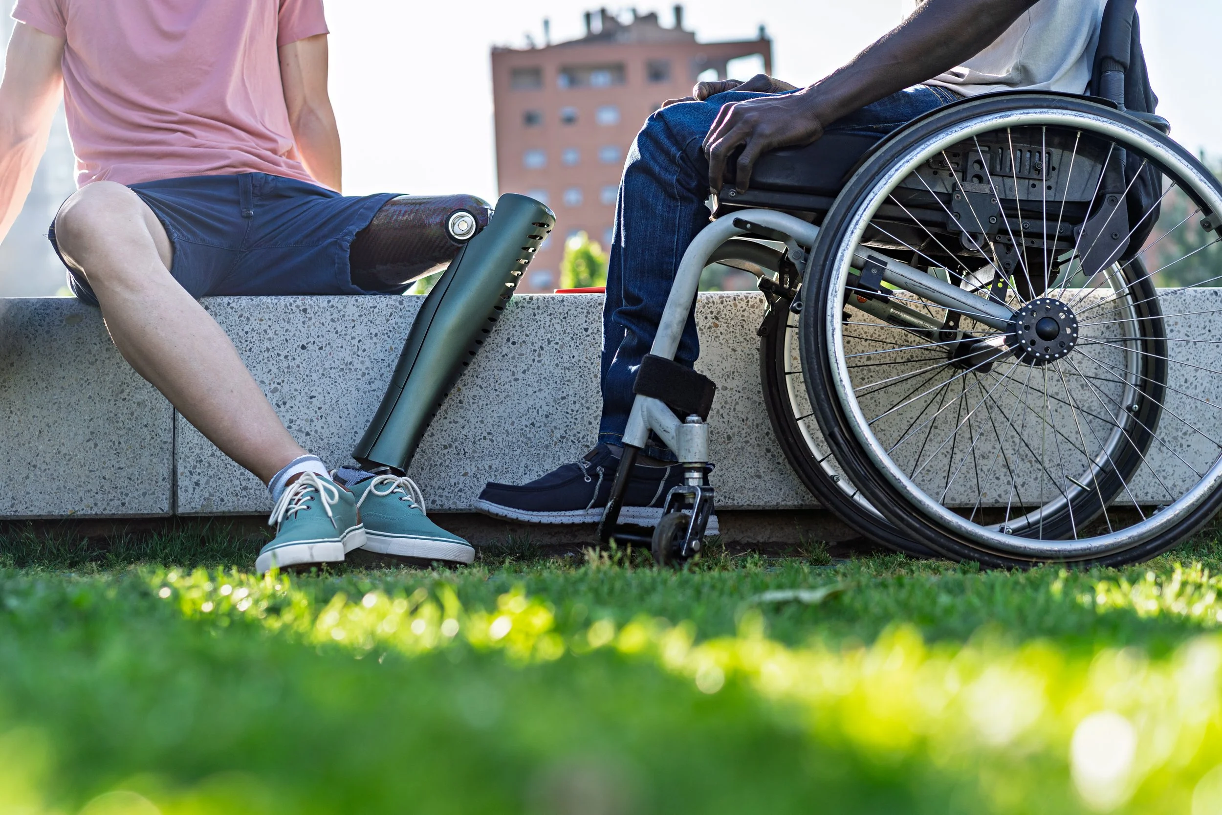 A person with a prosthetic leg sitting on a ledge next to a person in a wheelchair, outdoors on a sunny day with a building in the background.