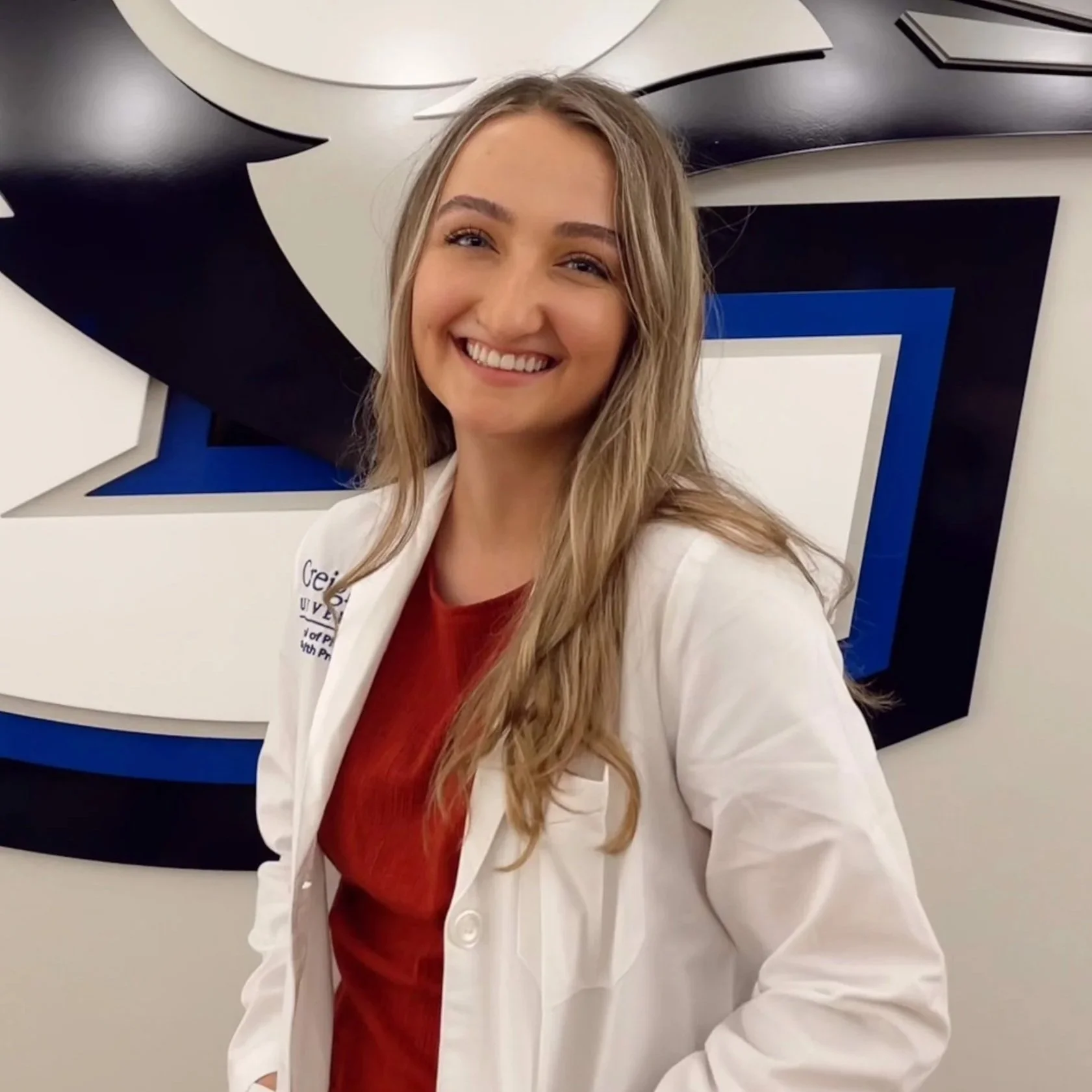 A smiling woman with long blonde hair wearing a white lab coat and a red shirt stands in front of a geometric black, white, and blue background.