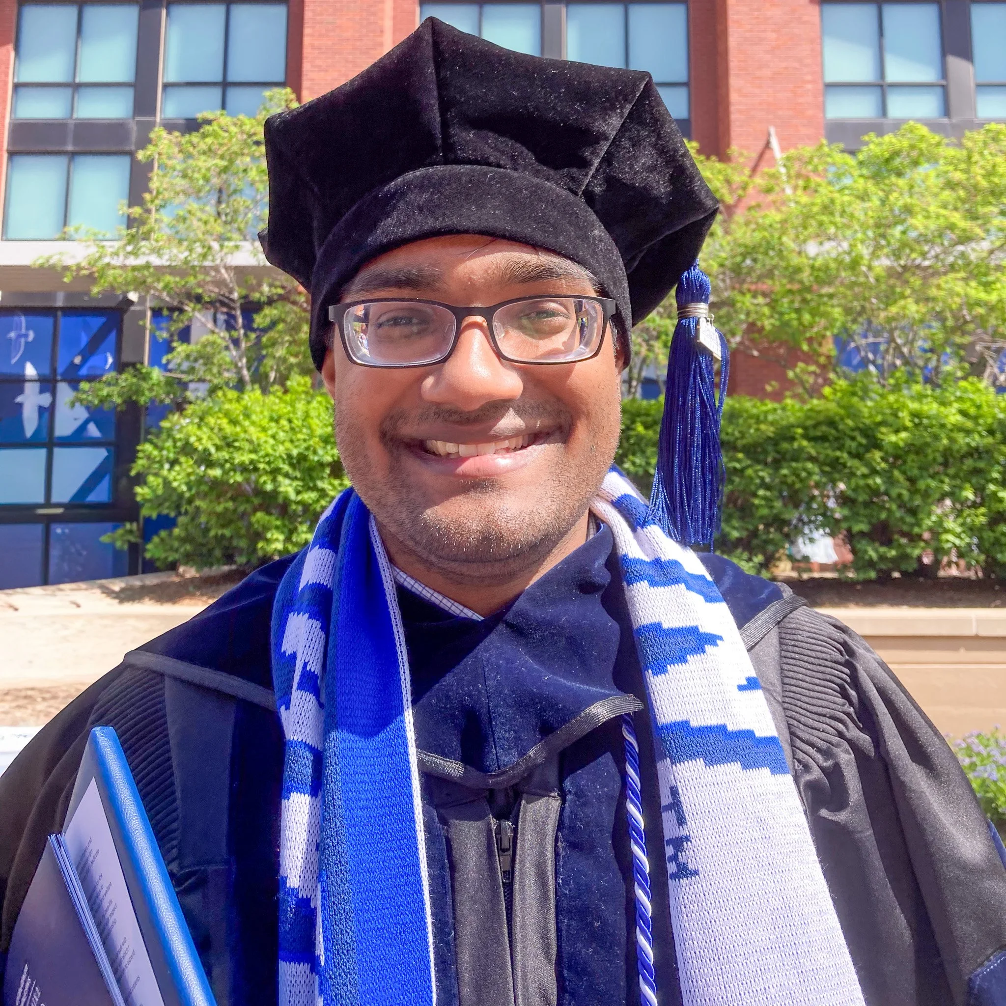 Close-up of a young man wearing glasses and a black graduation cap with a blue and white tassel, smiling outdoors in front of a modern brick and glass building with greenery.