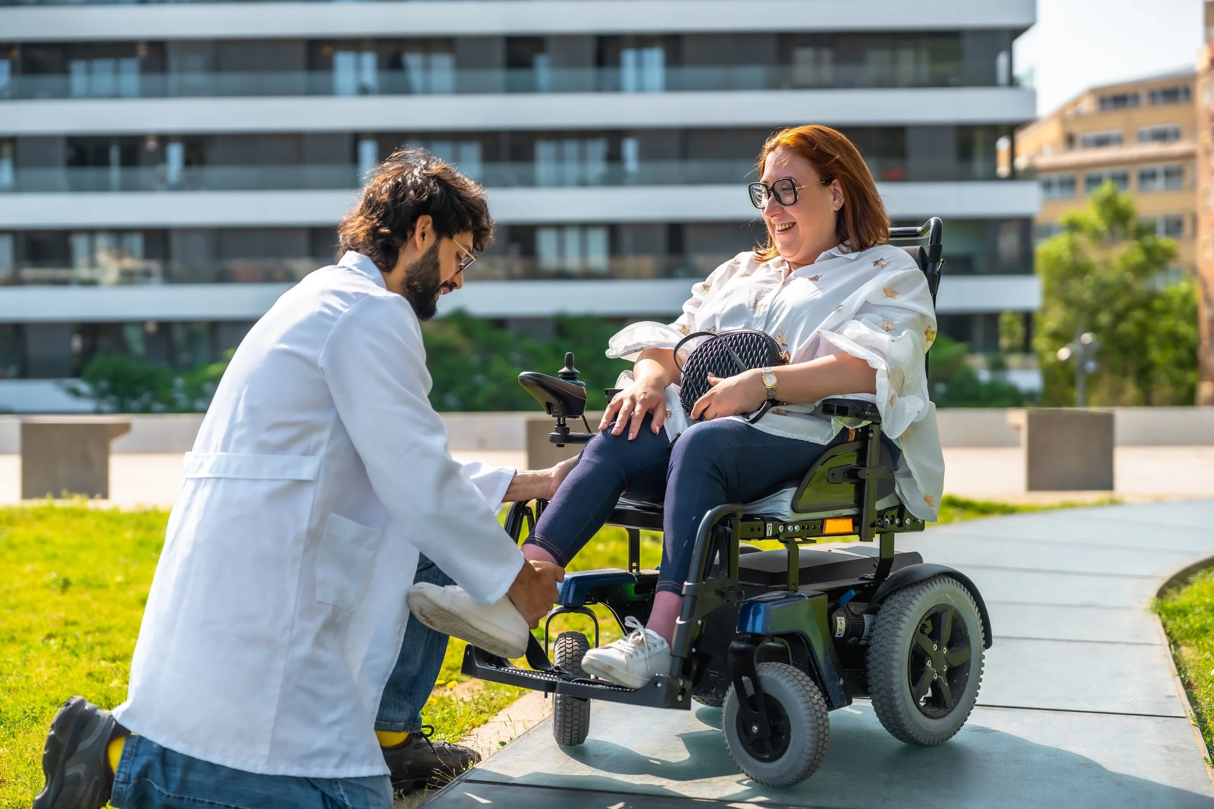 A woman in a wheelchair being assisted by a man in a white coat in an outdoor urban setting with modern building in the background.