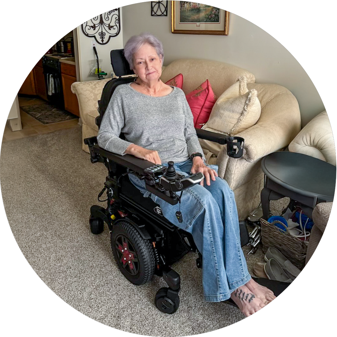 An elderly woman with short gray hair sitting in a motorized wheelchair in a living room with beige carpet, pillows, and a framed picture on the wall.