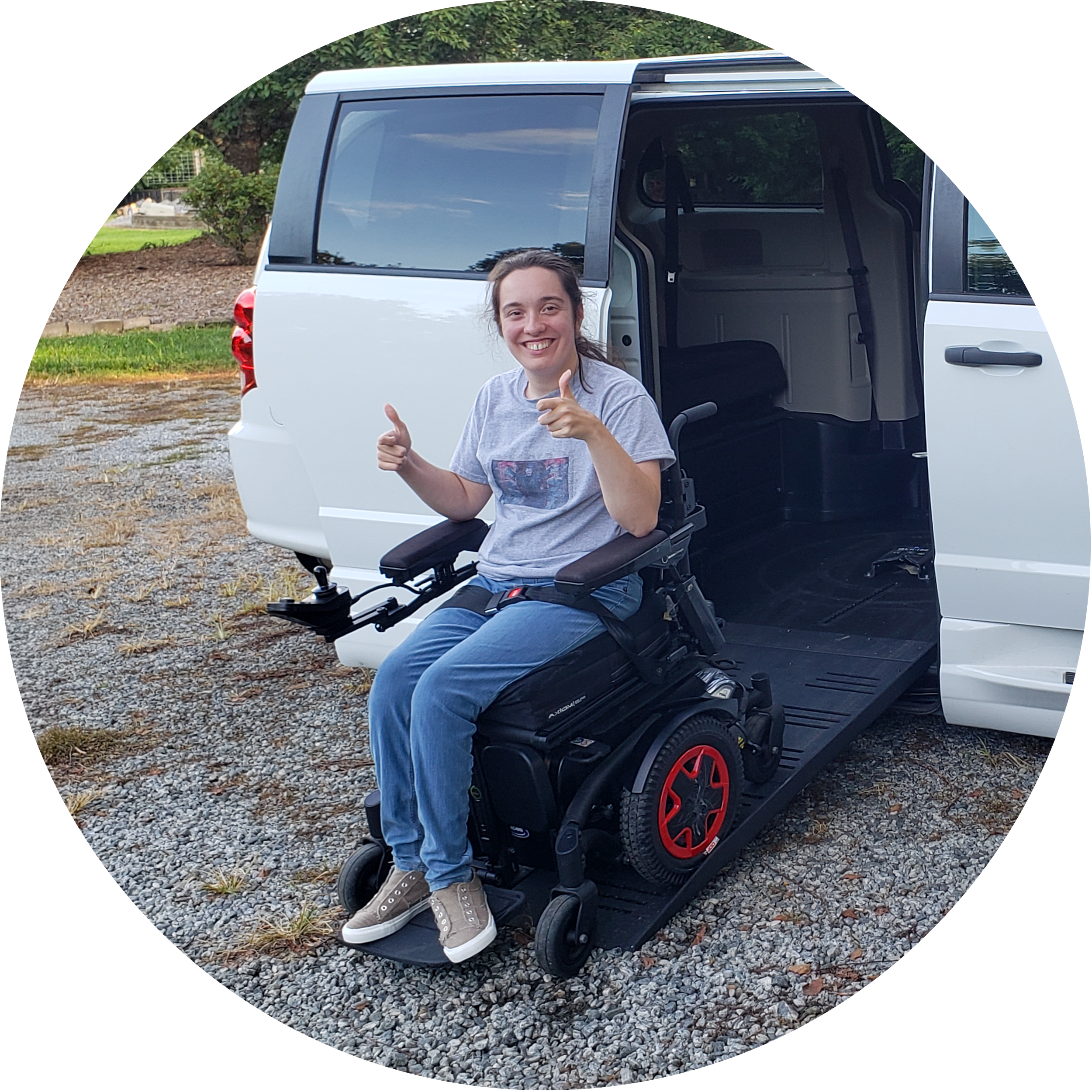 A woman sitting in a motorized wheelchair in front of an open van door, smiling and giving thumbs up, with green trees and bushes in the background.