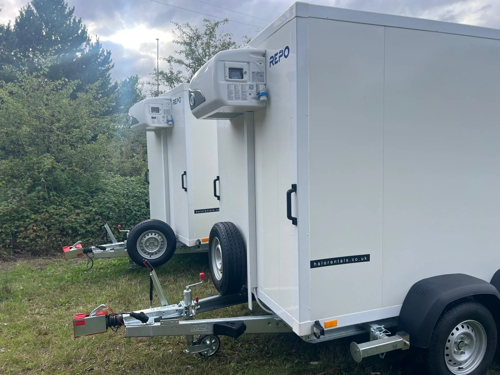 A white refrigerated or freezer trailer with two attached units on a grassy area, surrounded by green trees and a cloudy sky.