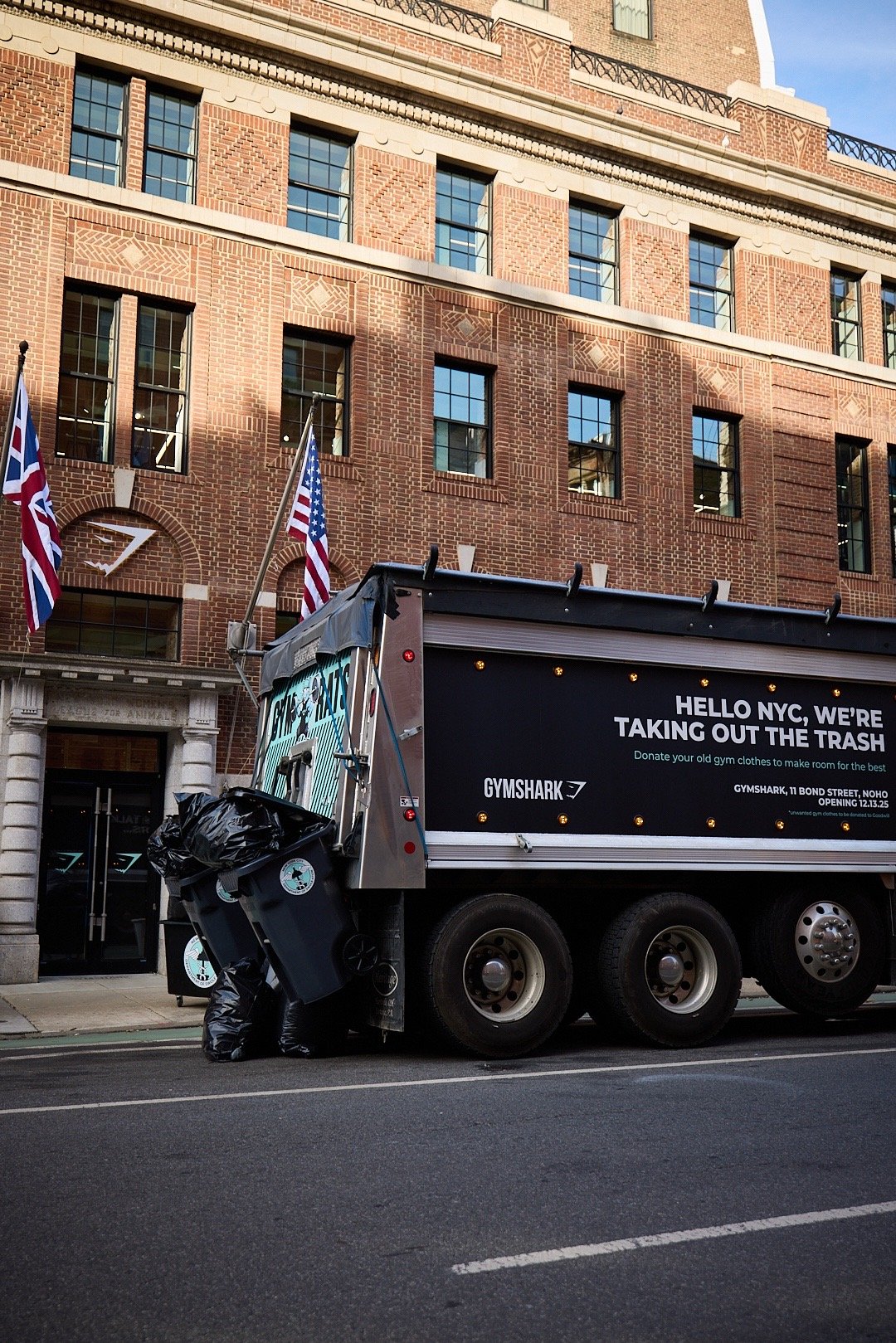 A garbage truck with a sign that says, 'HELLO NYC, WE'RE TAKING OUT THE TRASH,' parked on a city street in front of Gymshark NYC store.