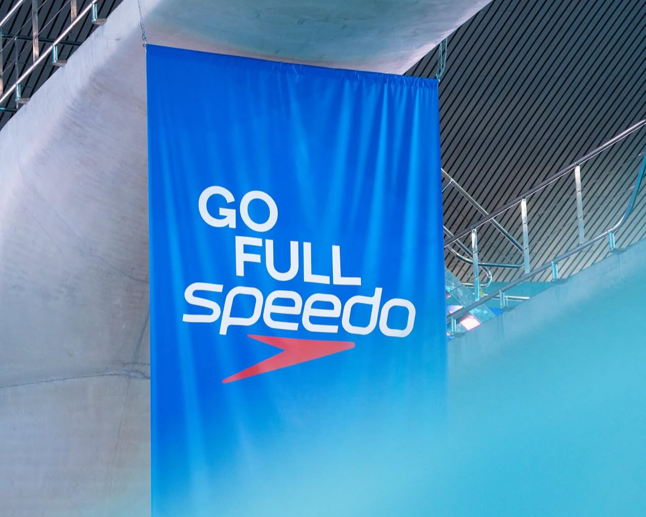 Blue banner with the words 'GO FULL speedo' and a red speed logo hanging in an indoor aquatic facility.
