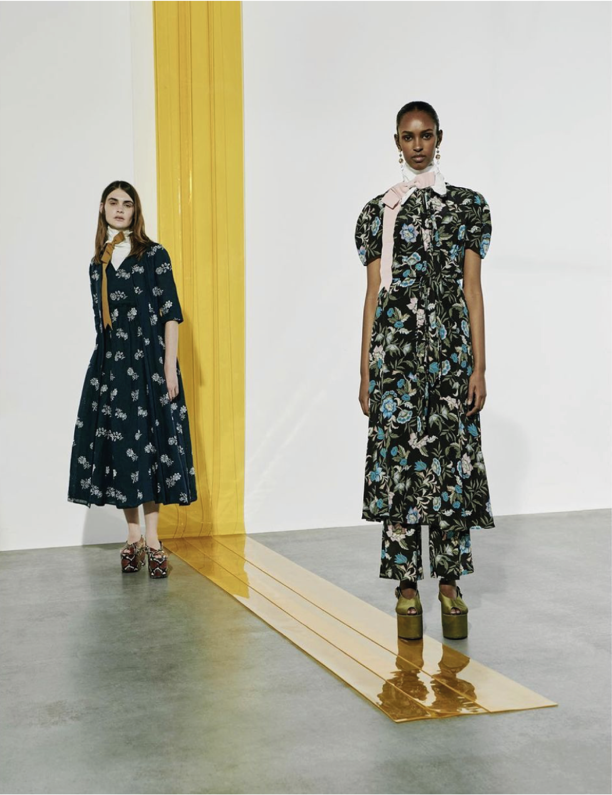 Two women standing in a minimalist indoor setting with a reflective golden floor. The woman on the left has long brown hair, wearing a dark blue floral dress and floral high heels. The woman on the right has dark skin, wearing a black floral dress wi