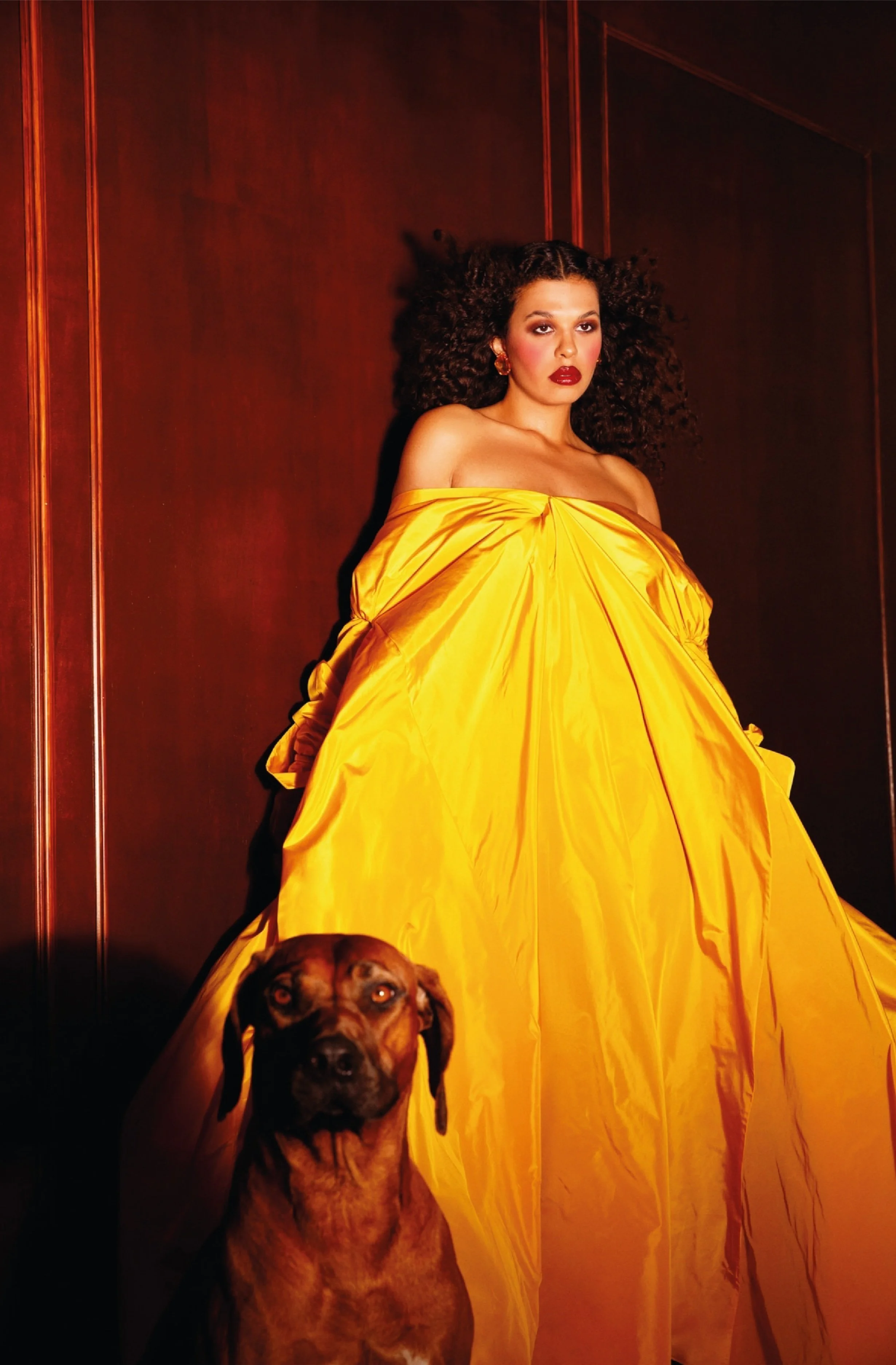 A woman with curly hair wearing a yellow off-the-shoulder gown and dark lipstick standing against a dark wooden wall, accompanied by a large dog sitting beside her.