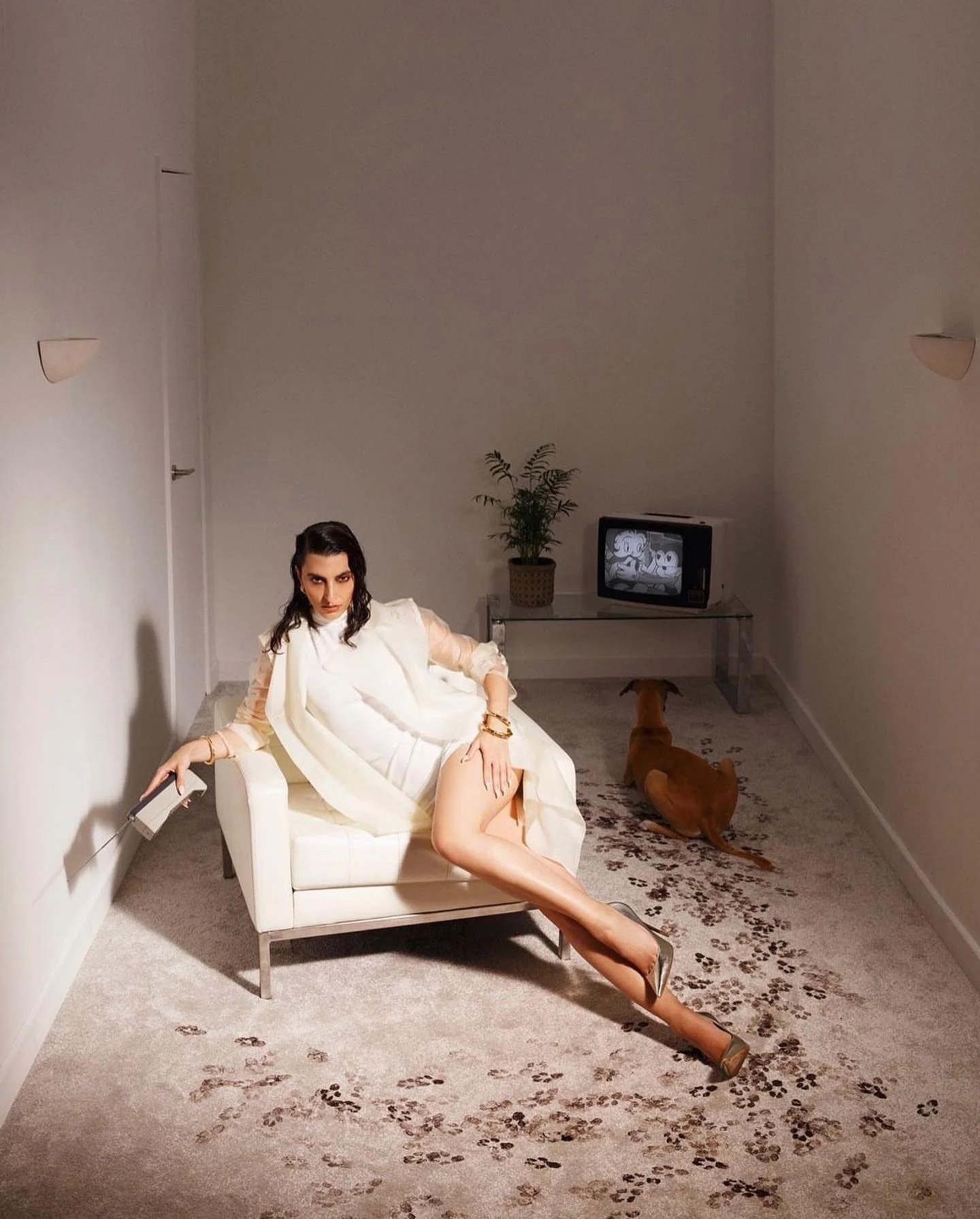 A woman dressed in white, sitting on a small white couch in a sparsely decorated room with a beige carpet. She has dark hair and a confident expression, with one leg crossed over the other and her left arm resting on the arm of the couch. A dog is lying on the floor nearby, and a TV, a potted plant, and a glass table are visible against the wall in the background.