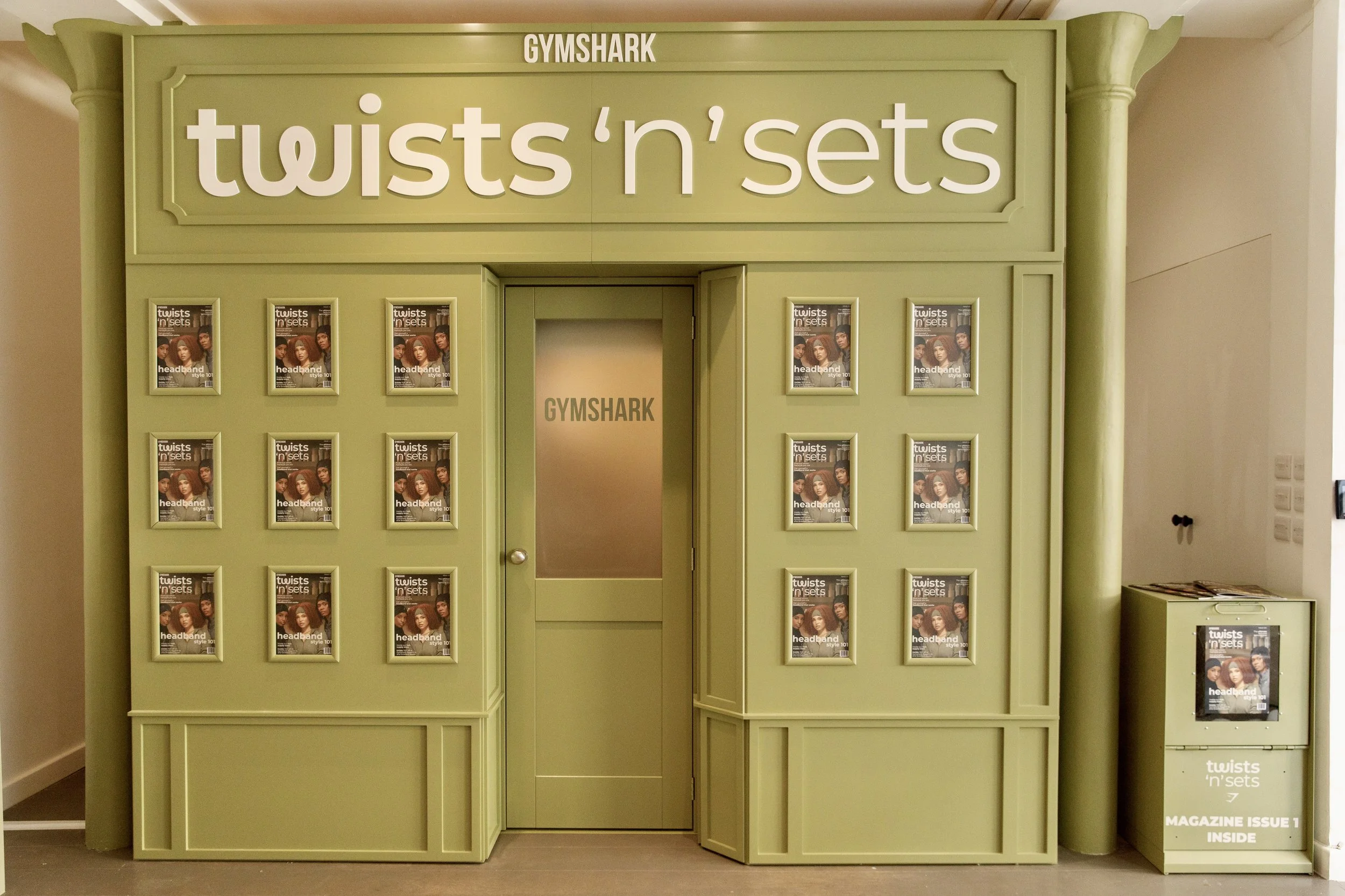 A green display kiosk with the signage 'twists 'n' sets' at the top, advertising headbands. The front features multiple magazine covers with models wearing various headbands. There is a door with the 'GYMSHARK' logo in the center and a small magazine stand at the bottom right.