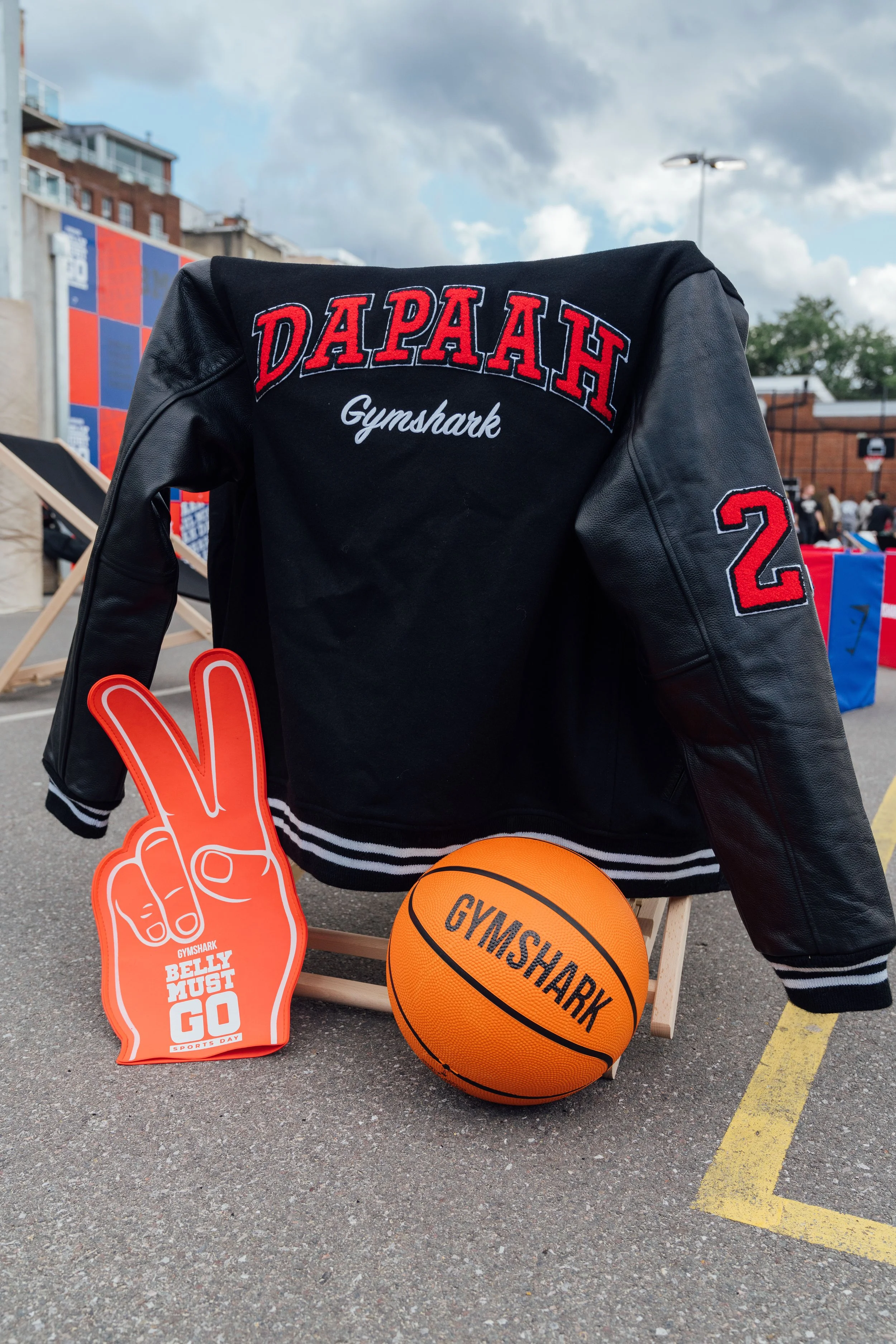 A black varsity jacket with red and white embroidery reading 'DAPA' and 'Gymshark,' a patch number 22, an orange Gymshark basketball, a foam hand with a peace sign and the text 'Belly Must Go Sports Day,' and a partial view of a foam finger.