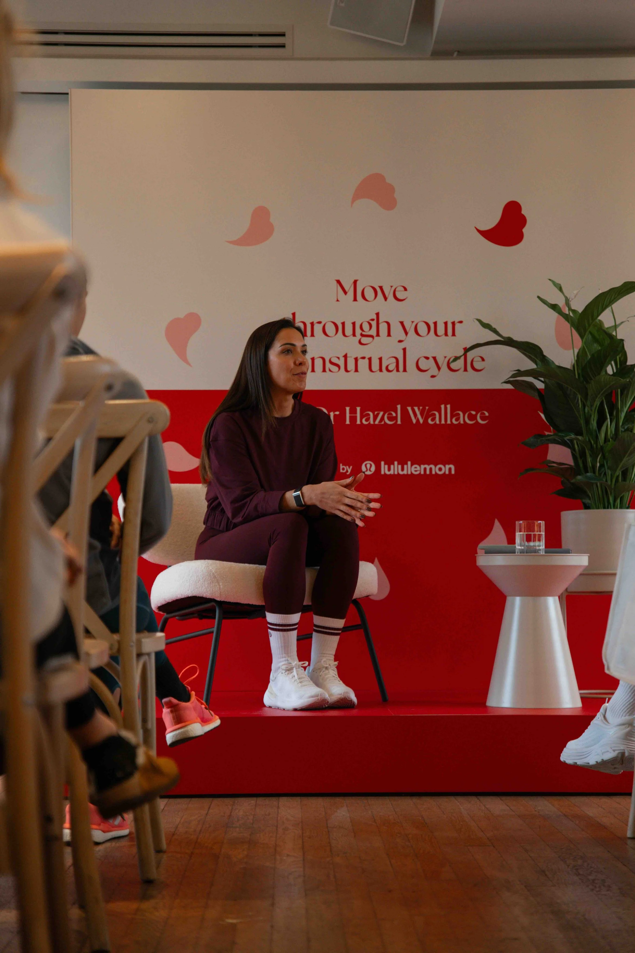 A woman seated on a stage, speaking during a presentation at an event about menstrual cycles, with a sign behind her that reads 'Move through your menstrual cycle' and a plant is visible to her right.