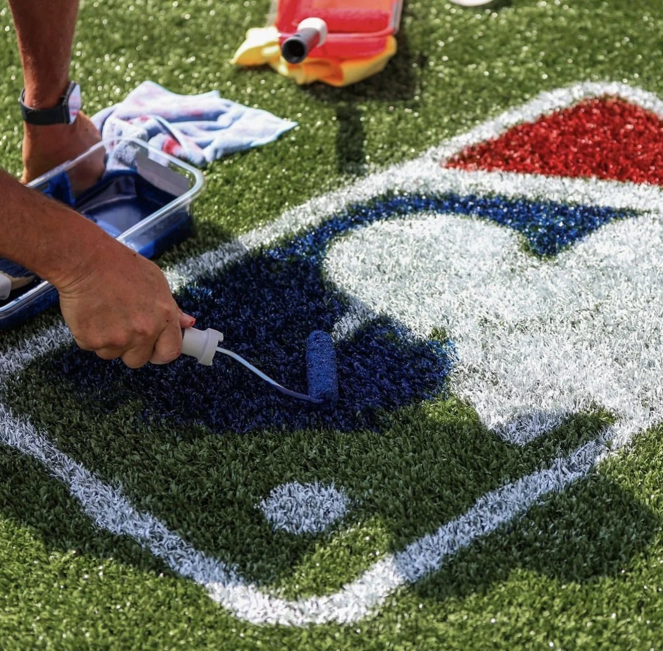 Person using a small roller to apply paint to a large artwork of a Toronto Blue Jays logo on a grassy surface.
