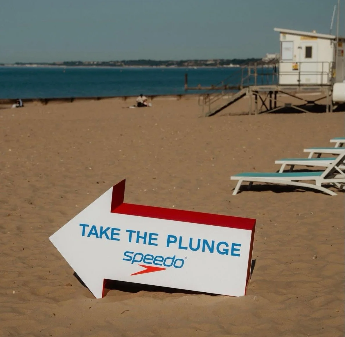 Beach scene with lounge chairs, a lifeguard station, and a sign with an arrow that says 'Take The Plunge' with the Speedo logo.