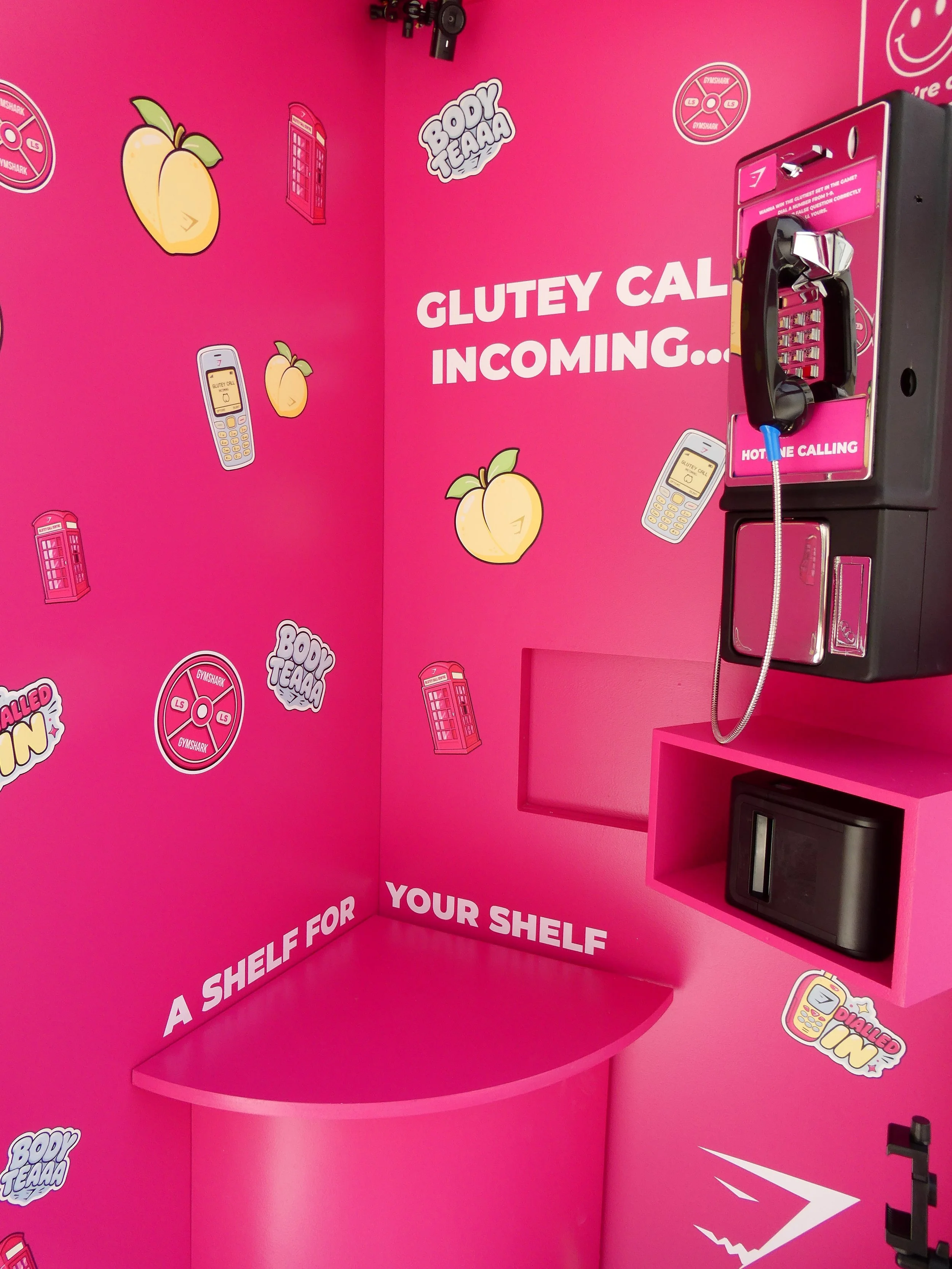Pink wall with stickers of peach emojis, red telephone booths, phones with the words 'GLUTEY CALL' on the screens, and speech bubbles saying 'BOY TEAAA' and 'DIALED IN'. There is a payphone mounted on the wall with a pink label that says 'HOTLINE CALLING'. There is a curved pink shelf attached to the wall.