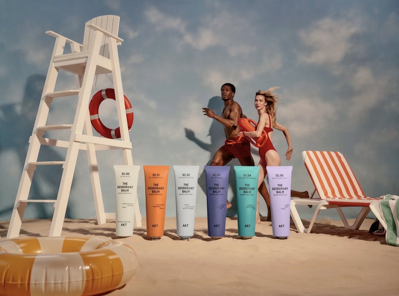 A beach scene features two people running along the sand, one man and one woman, with a lifeguard chair, a life preserver, two lounge chairs with orange and white stripes, and a set of colorful tubes of deodorant balm in the foreground.