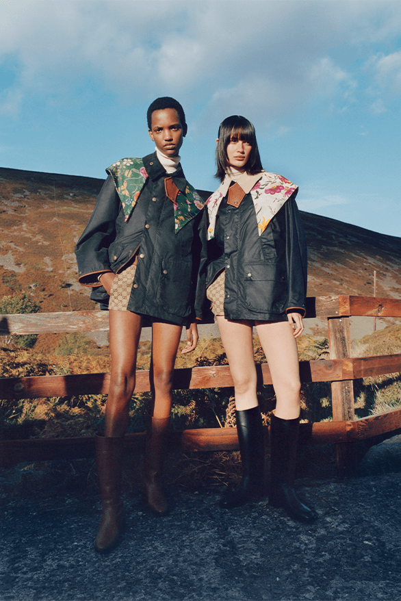 Two young women standing outdoors on a fence, wearing fashion jackets with floral shoulder details, one with tall boots, against a landscape with a hill and cloudy sky.