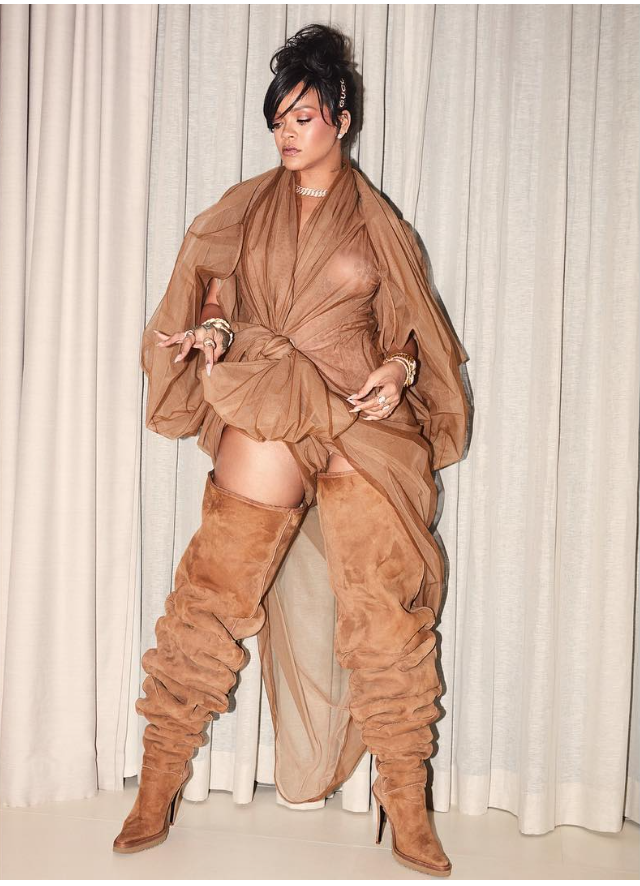 A woman with dark hair styled in an updo, wearing a brown sheer dress with large sleeves, thigh-high suede boots, and jewelry, standing in front of a curtain.