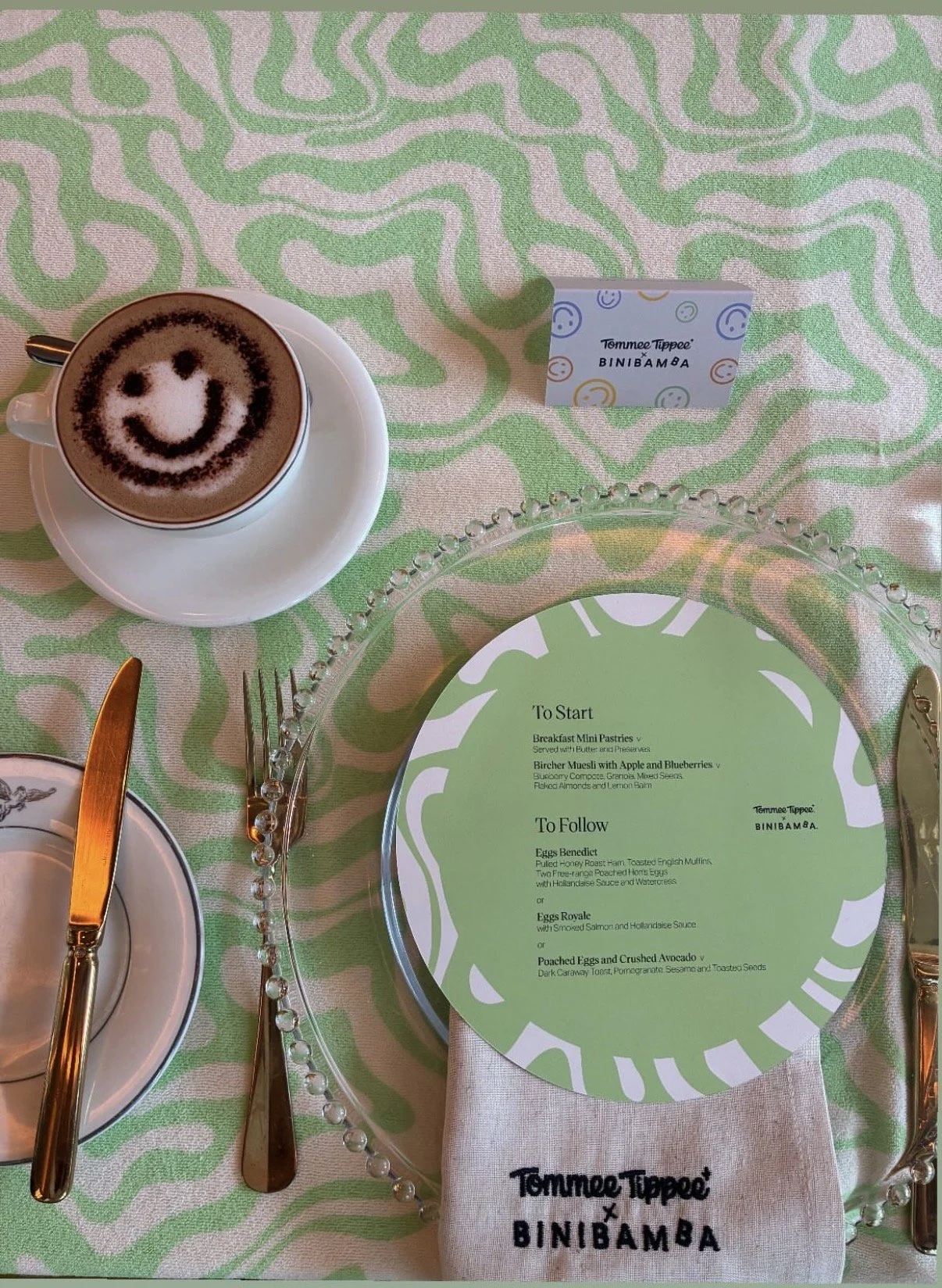A table setting with a cappuccino featuring a smiley face latte art, a cutlery set, a decorative glass charger plate with a menu, a patterned green and white tablecloth, and a small card that says "Tommee Tippee x BINIBAMBA".