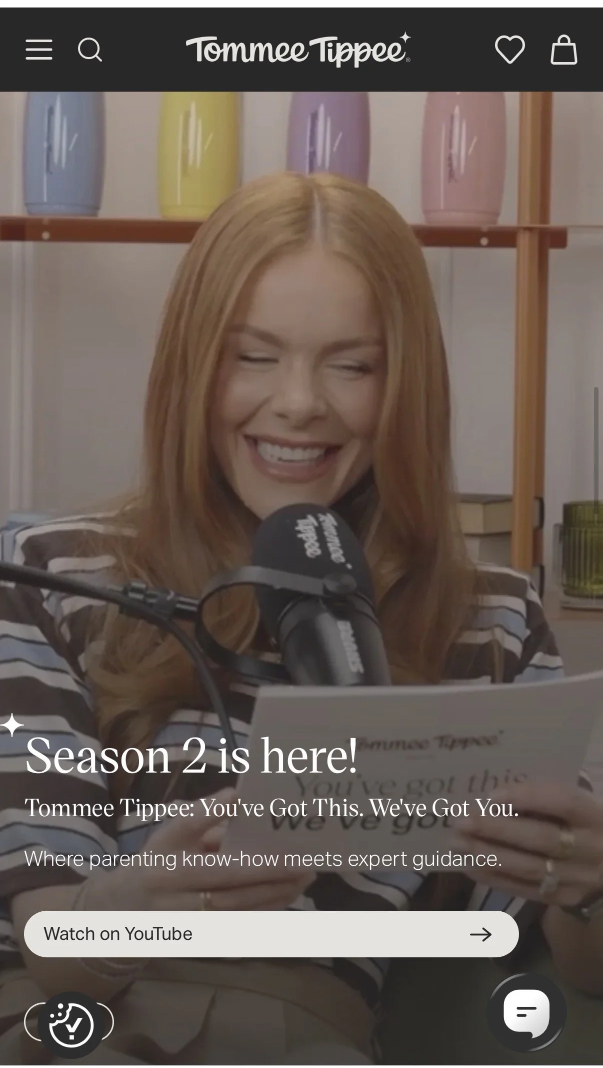 A woman with red hair smiling and speaking into a microphone, holding a piece of paper, with shelves of colorful vases behind her. The overlay text reads: "Season 2 is here!" and "Tommee Tippee: You've Got This. We've Got You."