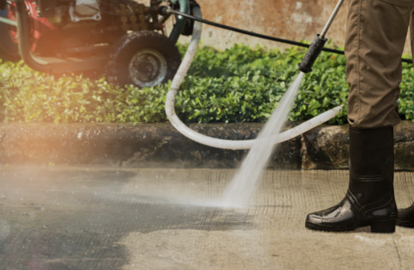Person wearing black rubber boots and brown pants pressure washing a concrete sidewalk.