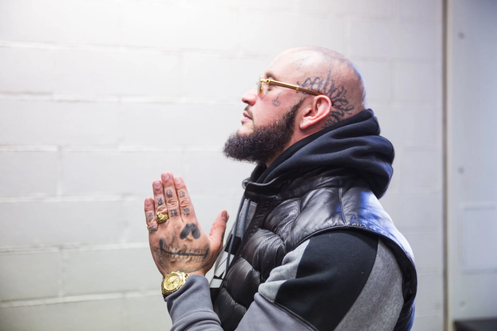 Homme tatoué en praying position, portant des lunettes dorées, un manteau en cuir et une montre dorée.