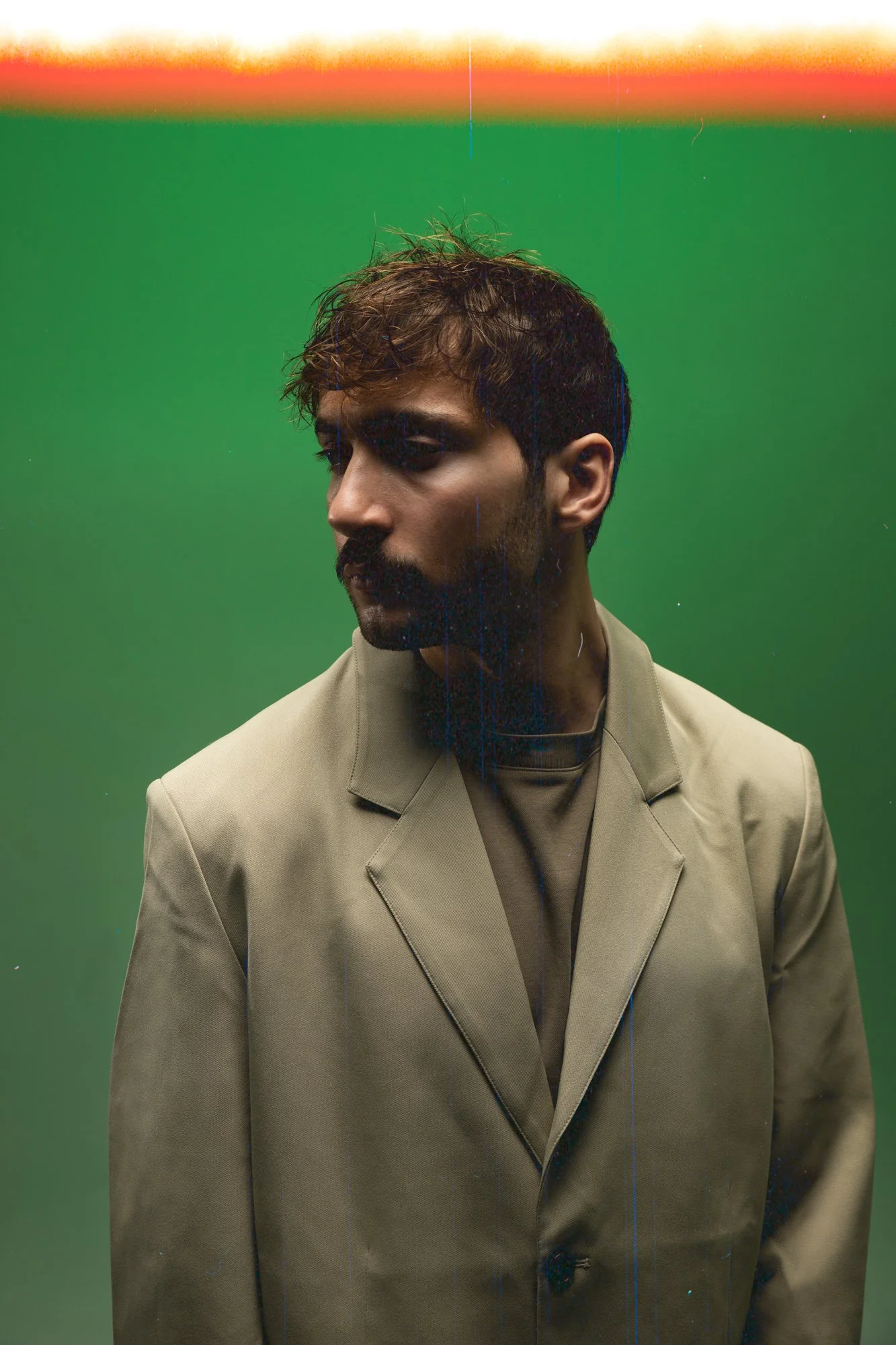 Portrait d'un homme à la coupe de cheveux bouclée portant un costume beige, regardant vers la gauche, avec un fond vert et un dégradé de couleurs chaudes en haut.