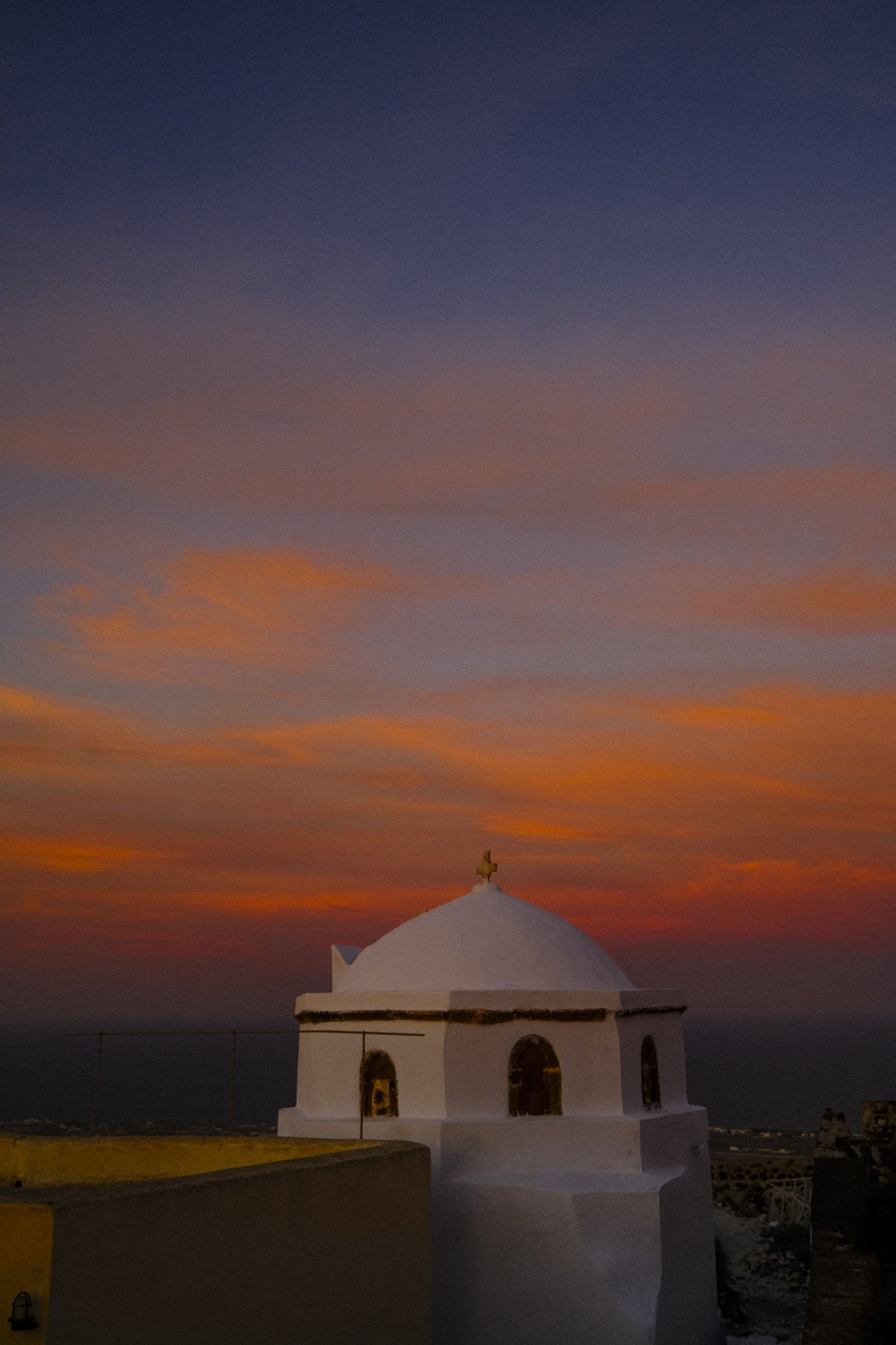 Une petite chapelle blanche avec un dôme et une croix au sommet, sous un ciel au coucher du soleil avec des couleurs orange, rose et violet à santorin