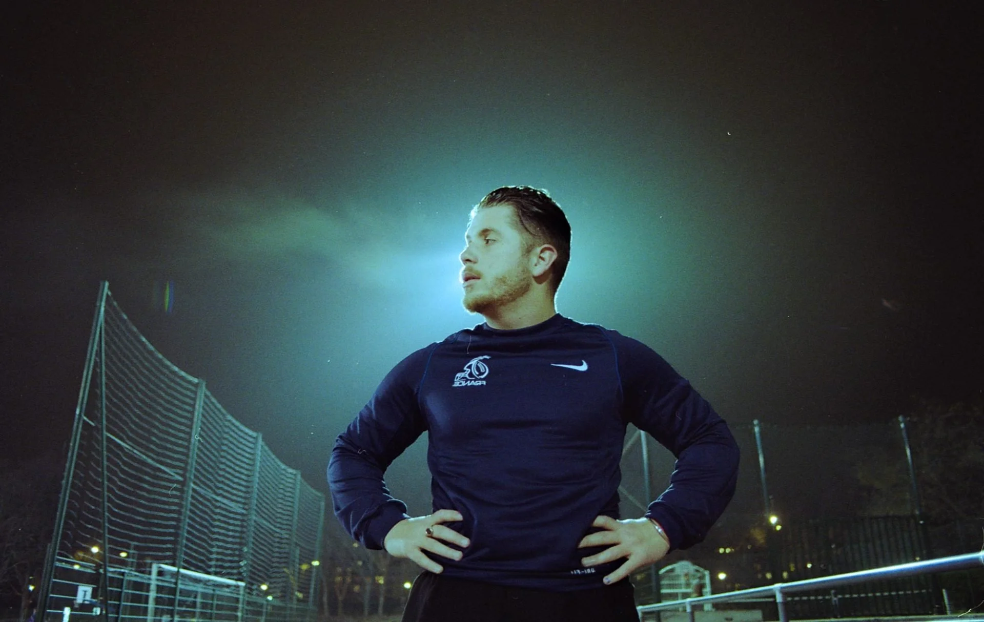 Paul Deby entraîné dans un terrain de sport en pleine nuit, portant un t-shirt noir Nike avec un logo blanc, éclairé par une lumière derrière lui et entouré d'une clôture de sport.