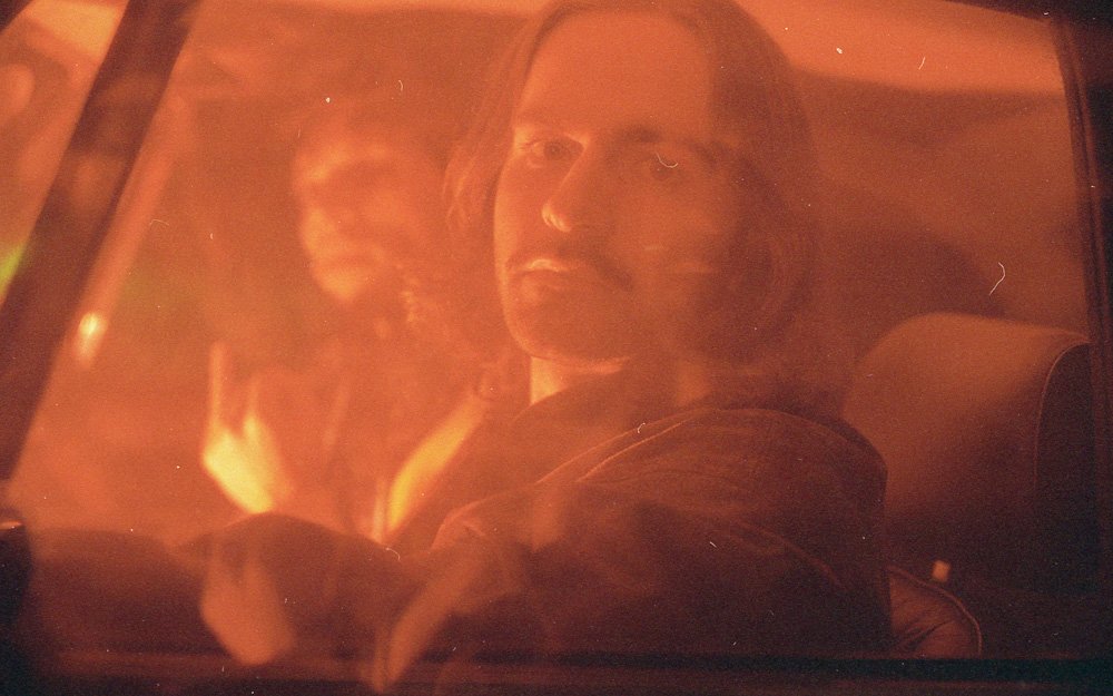 Une scène de film avec deux personnes dans une voiture, éclairée par une lumière orange ou rouge.
