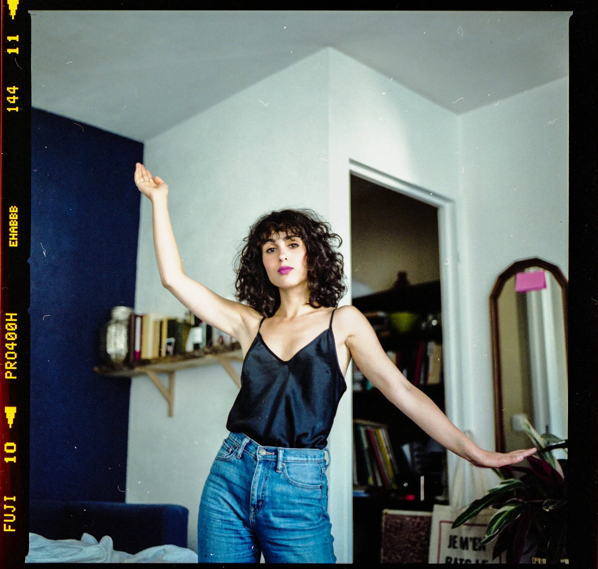 Une femme avec des cheveux bouclés, portant un haut en satin noir et un jean, pose dans une pièce avec livres et décorations derrière elle.